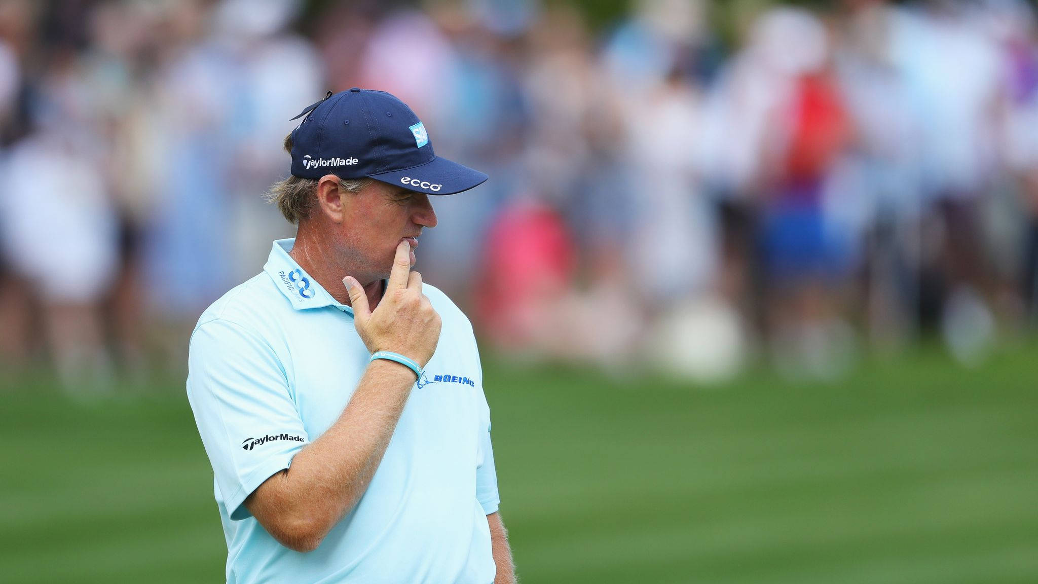 Ernie Els 向右凝視 壁紙 — 一位專注的高爾夫球手，身穿淺藍色襯衫和海軍藍帽子，在陽光明媚的果嶺上思考下一桿，身後有模糊的觀眾。