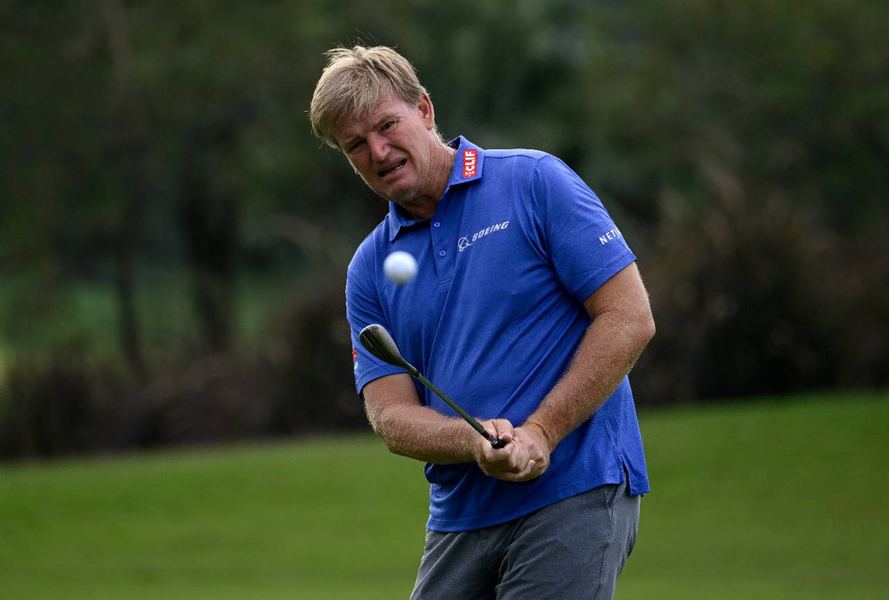 Ernie Els 高爾夫揮杆 壁紙 — 一位身穿藍色襯衫的高爾夫球手，在模糊的綠色背景下，專注地揮桿，球在空中飛過。
