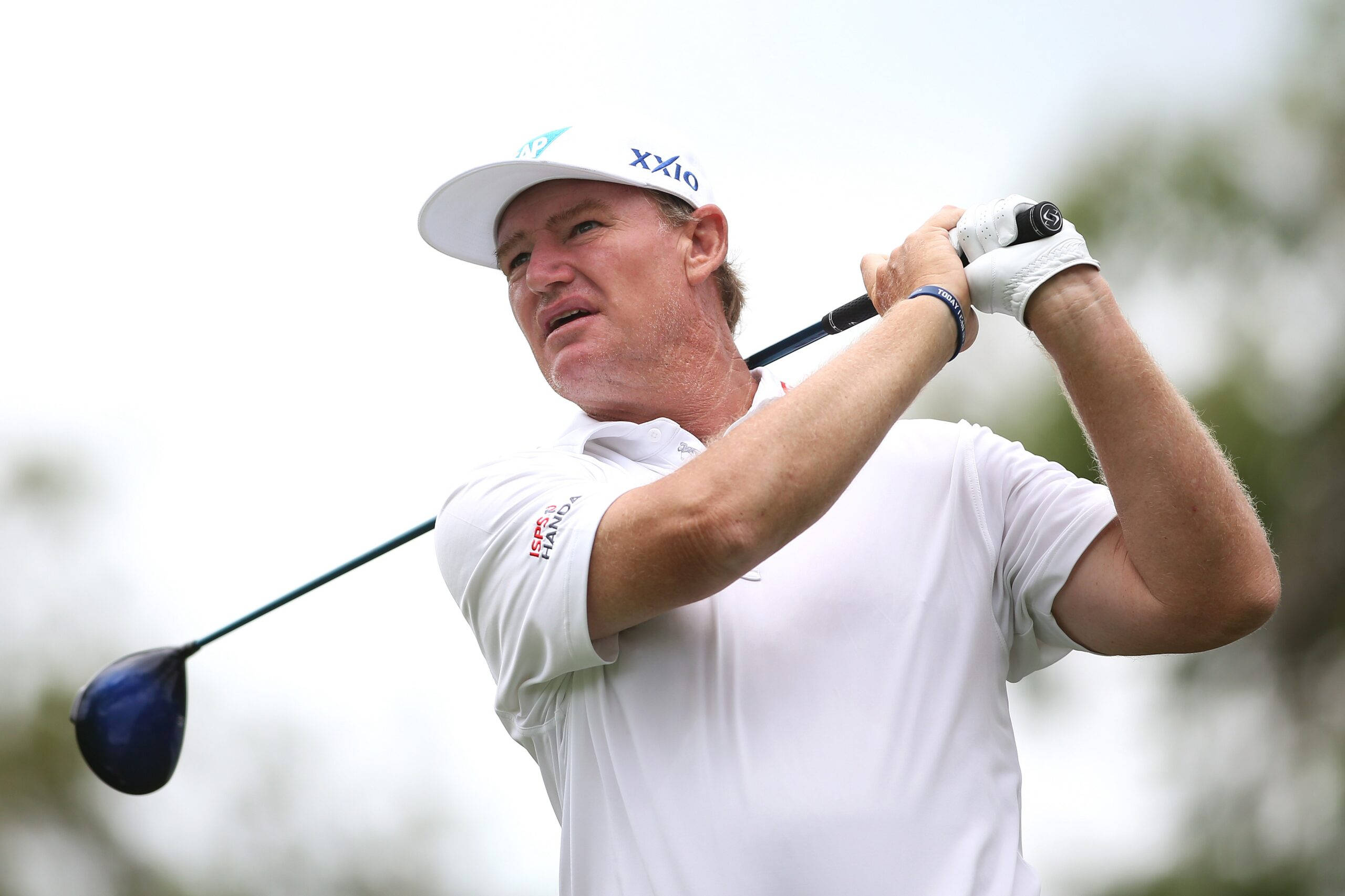 Ernie Els 身穿白色服裝 壁紙 — 一位身穿白色襯衫和帽子的男性高爾夫球手，在明亮的戶外背景下，專注地揮動一號木桿。