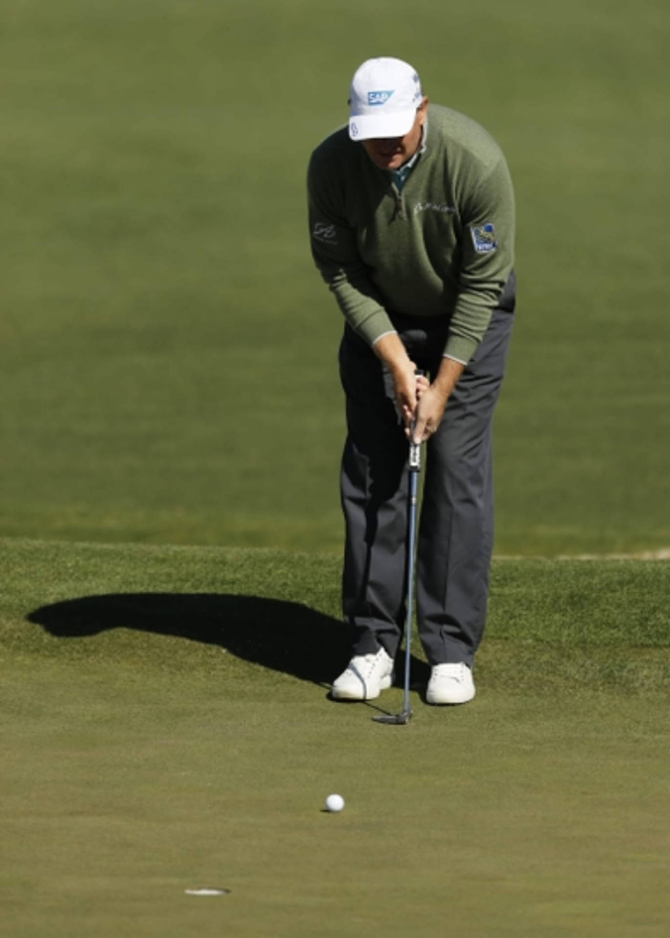 Ernie Els 推桿瞬間 壁紙 — 一位身穿綠色毛衣、頭戴白色帽子的高爾夫球手，在陽光普照的果嶺上專注地瞄準推桿路線。