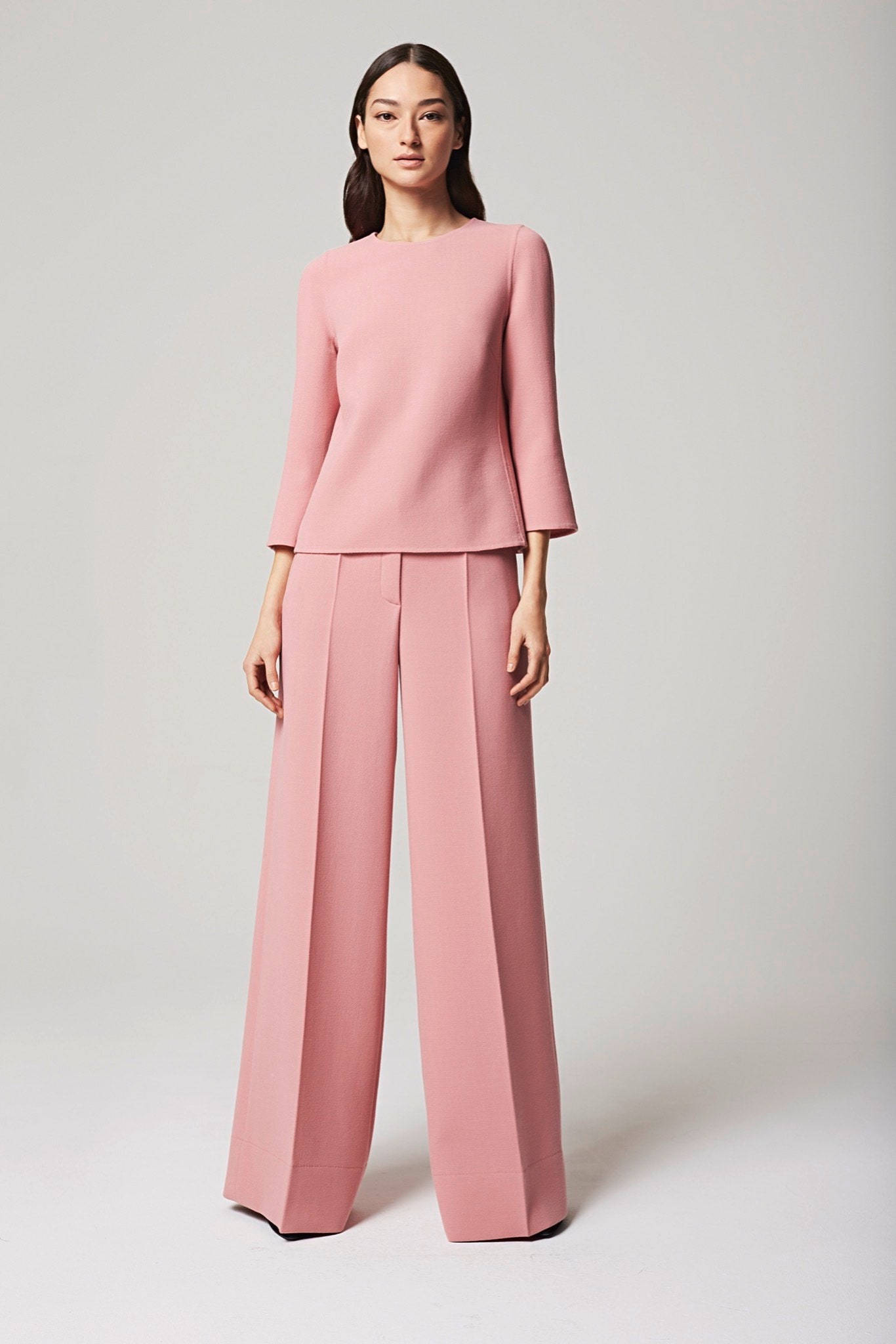 Escada 2016度假系列 壁紙 — 一位穿著柔和粉紅色訂製西裝褲套裝的優雅女性，站在簡約工作室背景前，光線柔和中性。