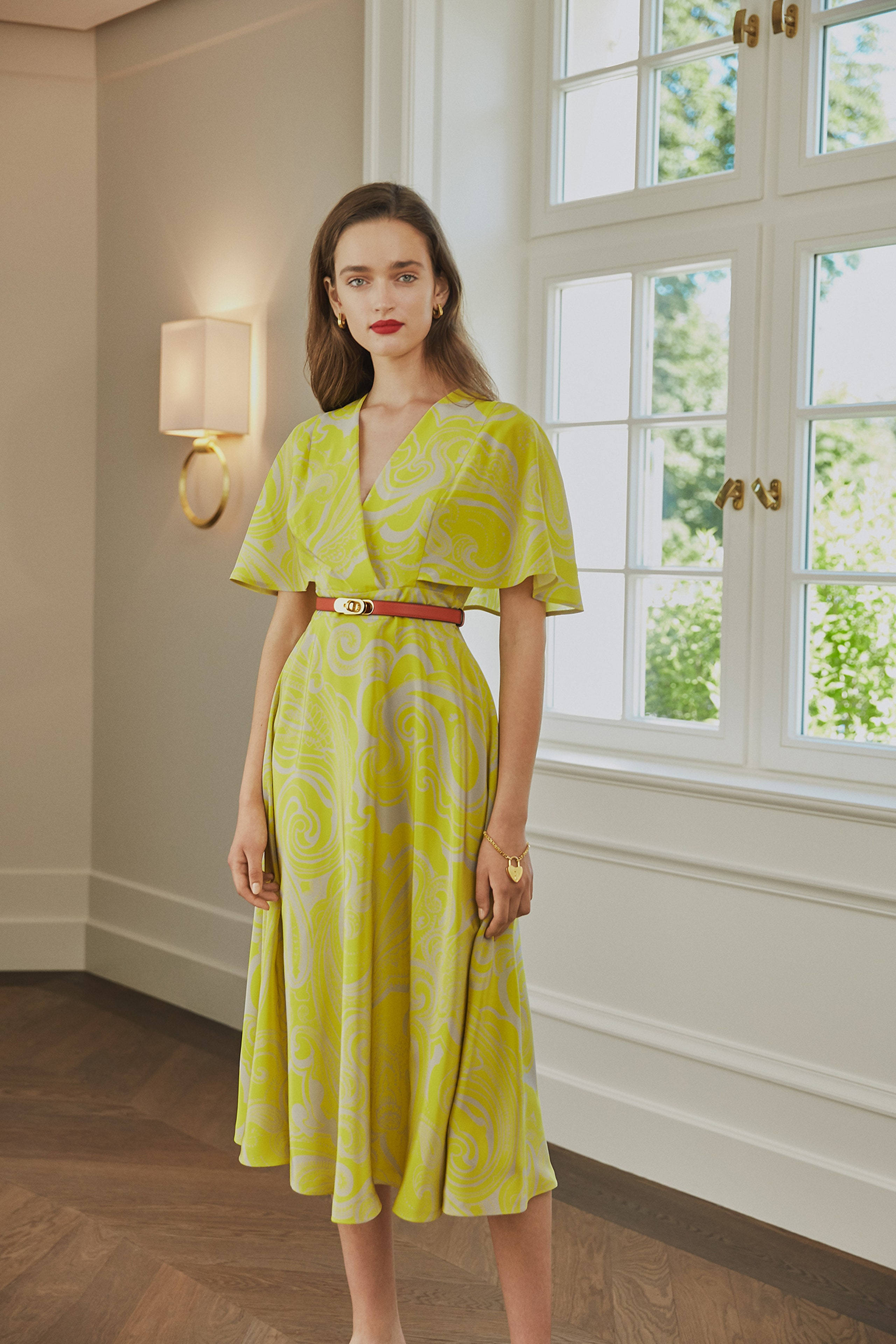 Escada 2020春季系列 壁紙 — 一位身穿鮮豔黃色圖案連衣裙、繫著紅色腰帶的女性，在簡約風格的室內明亮窗邊優雅地站立。