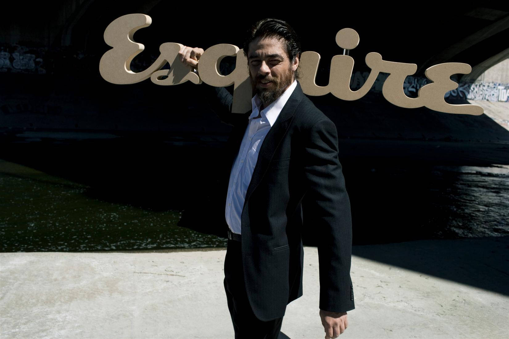 本尼西歐·德·托羅《Esquire》十月號（2007） 壁紙 — 一名身穿深色西裝的男子自信地站在一個大型 Esquire 標誌下，背景有塗鴉牆壁和水面。