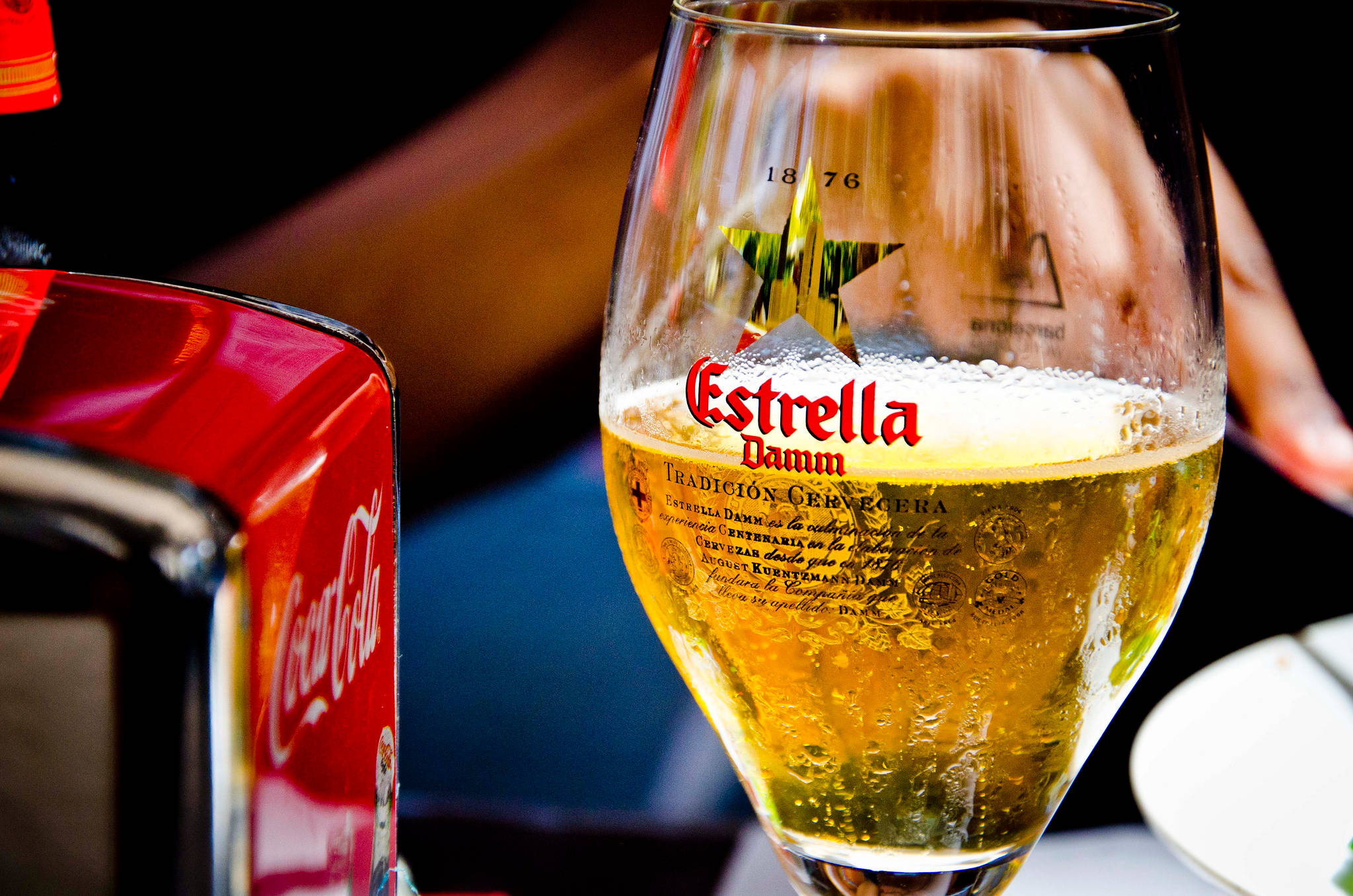 晶瑩的 Estrella Damm 啤酒杯 壁紙 — 一杯冰凍的 Estrella Damm 啤酒，帶有水珠，放在一瓶紅色可口可樂旁邊，環境燈光昏暗。