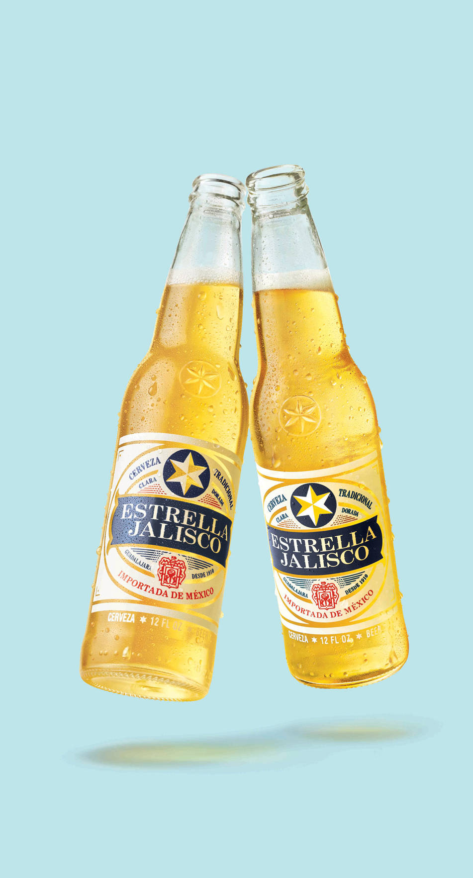 兩瓶Estrella Jalisco啤酒在藍色背景上碰撞，帶有冷凝水滴。