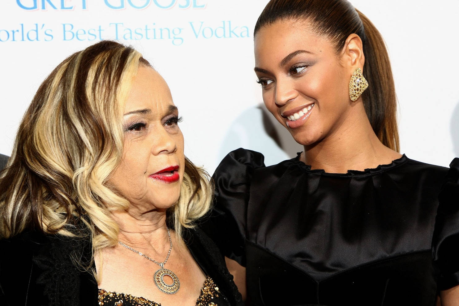 Etta James 與 Beyoncé 壁紙 — 兩位女性在品牌背景前熱情微笑，一位金色捲髮紅唇，另一位身穿黑色緞面禮服，佩戴珠寶耳環。