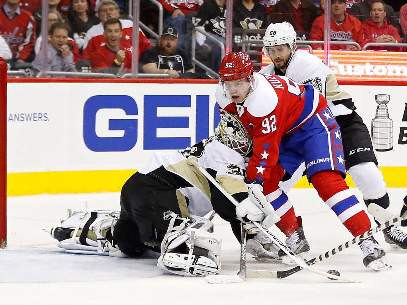 Evgeny Kuznetsov 動態賽場表現 壁紙 — 一名身穿黑色和金色的冰球守門員飛身撲救，試圖擋住射門，而一名紅藍球員在激烈的比賽中向前壓迫。