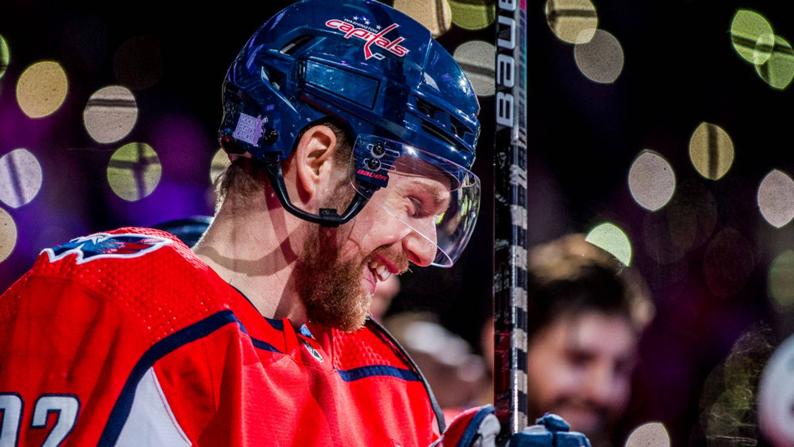 Evgeny Kuznetsov 展露迷人微笑 壁紙 — 一位微笑的曲棍球運動員身穿紅色Capitals球衣，在充滿活力的散景照明場館背景下握著球桿。
