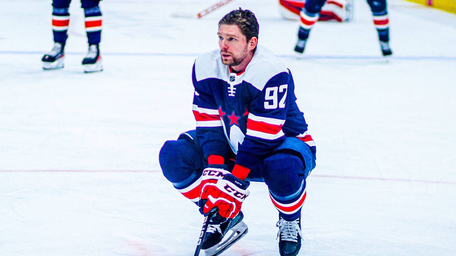 Evgeny Kuznetsov 場上休憩 壁紙 — 一名身穿藍、紅、白色球衣的冰球運動員在比賽中於冰場上激烈蹲下。