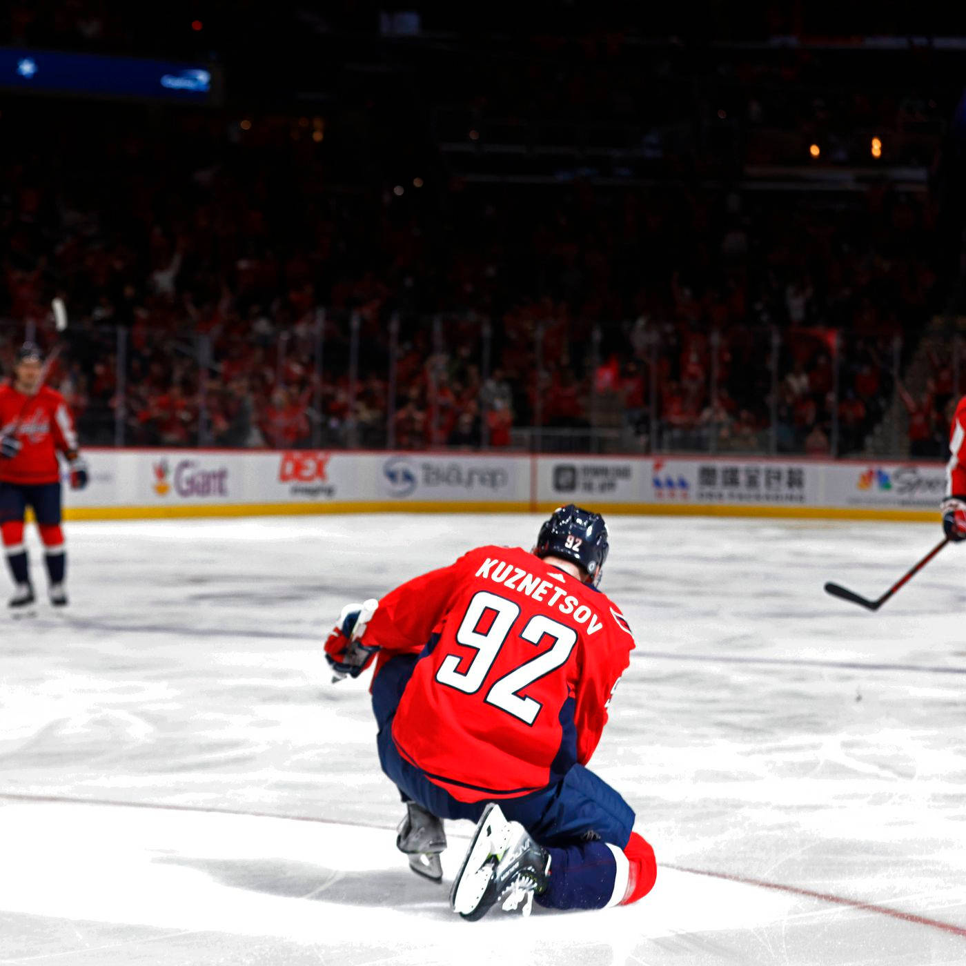 Evgeny Kuznetsov 後視角度 壁紙 — 一名身穿紅色92號球衣的冰球運動員在冰場上跪下，玻璃後面有模糊的人群。