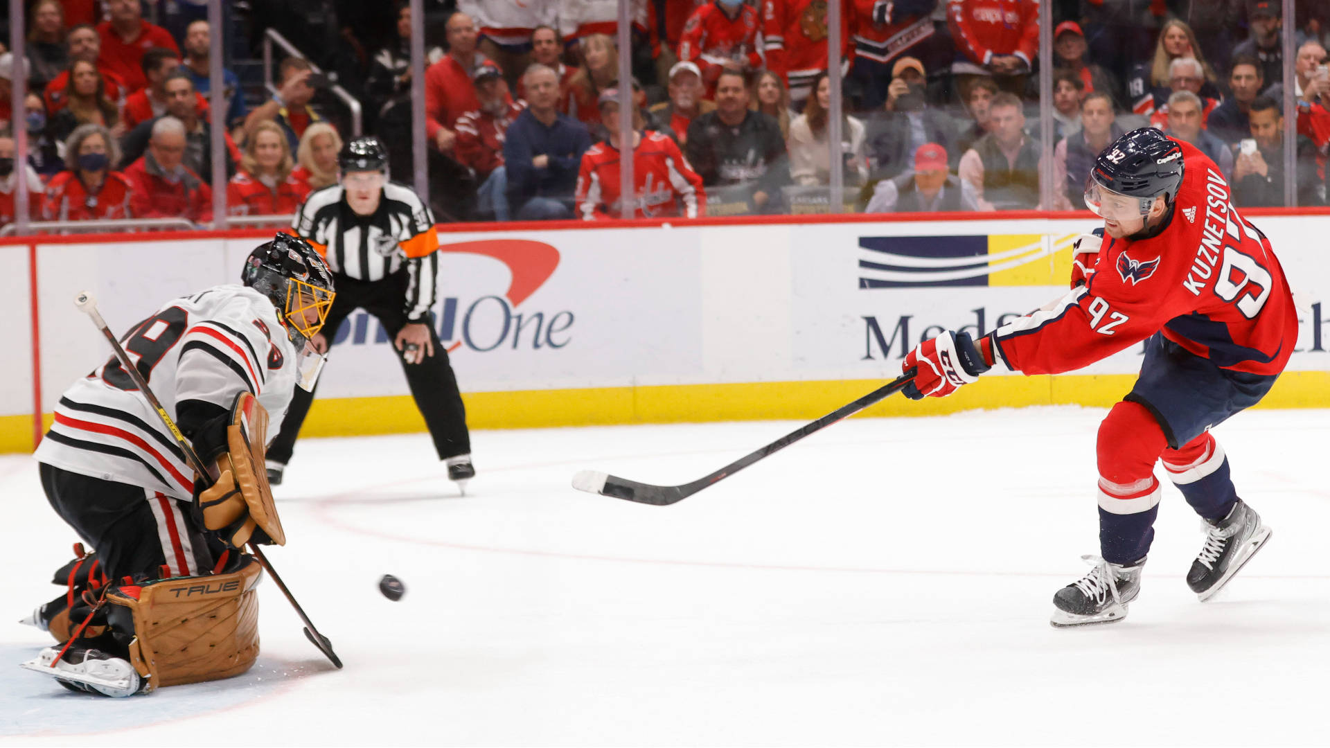Evgeny Kuznetsov 擊入制勝球 壁紙 — 一名身穿紅色球衣的冰球運動員射門，守門員飛身撲救，背景有觀眾和裁判。