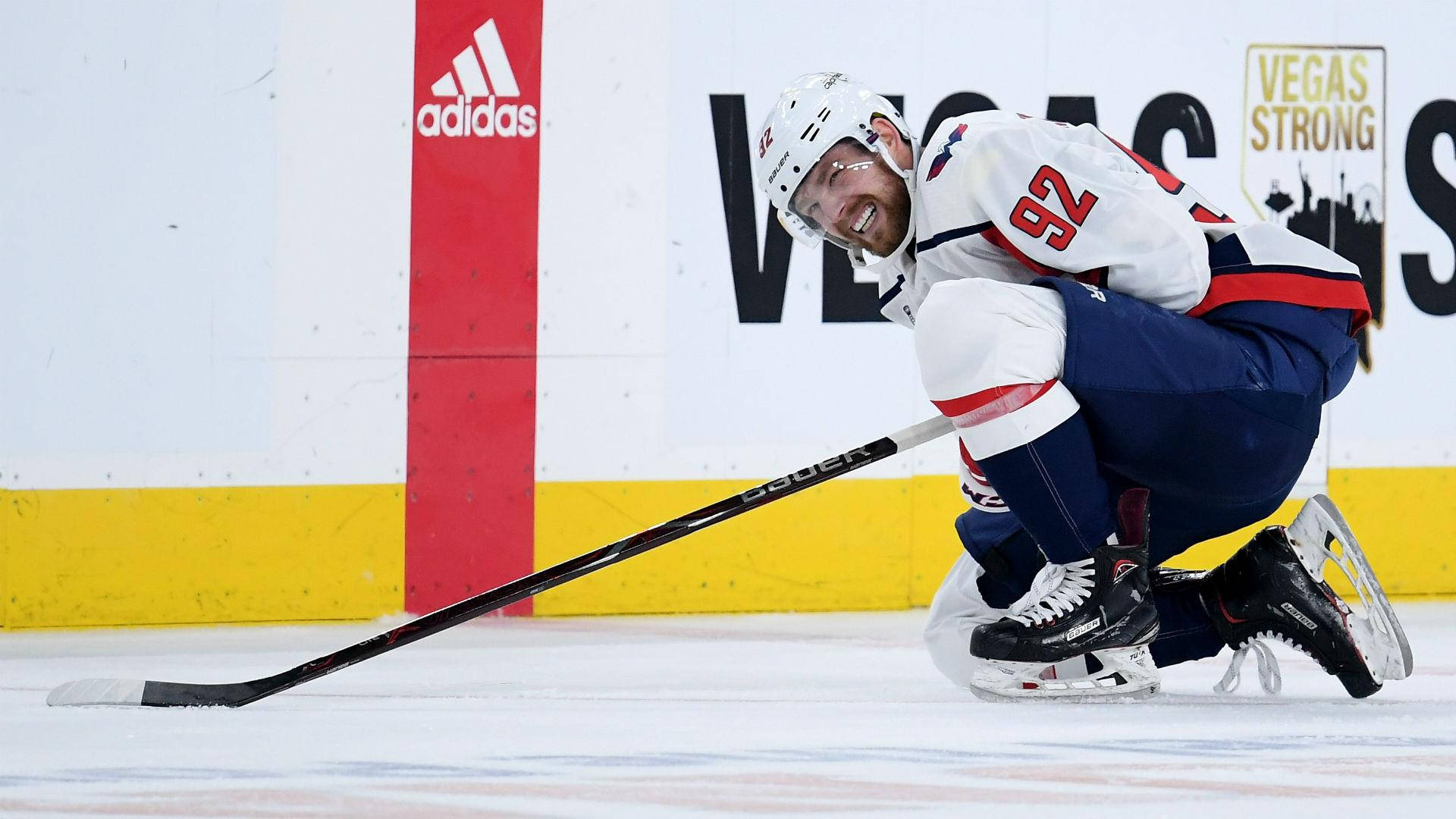 Evgeny Kuznetsov 場上受傷 壁紙 — 一名身穿白色92號球衣的冰球運動員在冰上跪著微笑，球桿放在身旁，旁邊有Vegas Strong橫幅。
