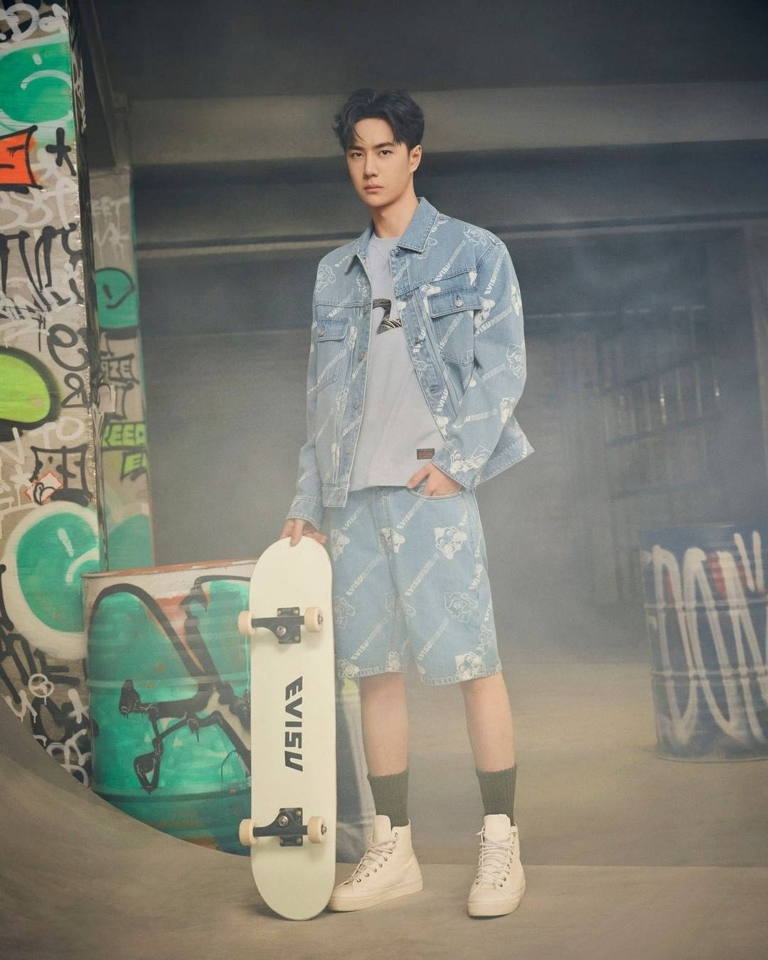 王一博 Evisu 滑板造型 壁紙 — 一位穿著圖案牛仔套裝的年輕男子自信地站在一個佈滿塗鴉的都市滑板公園中，手持白色滑板。