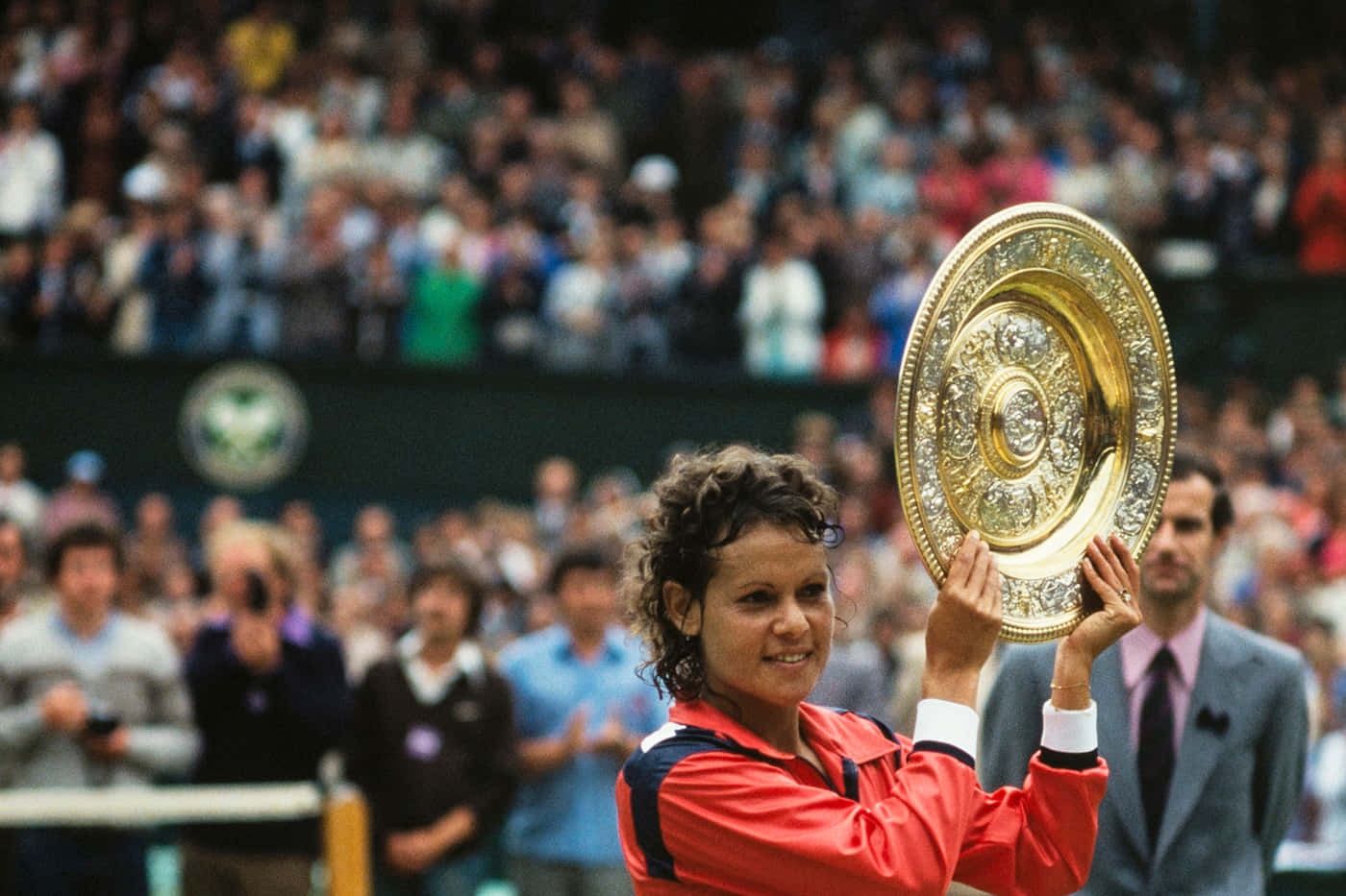 Evonne Goolagong Cawley 手捧溫布頓獎盃 壁紙 — 一位微笑的網球選手身穿紅色外套，在歡呼的人群面前自豪地高舉一個華麗的金色獎盃。