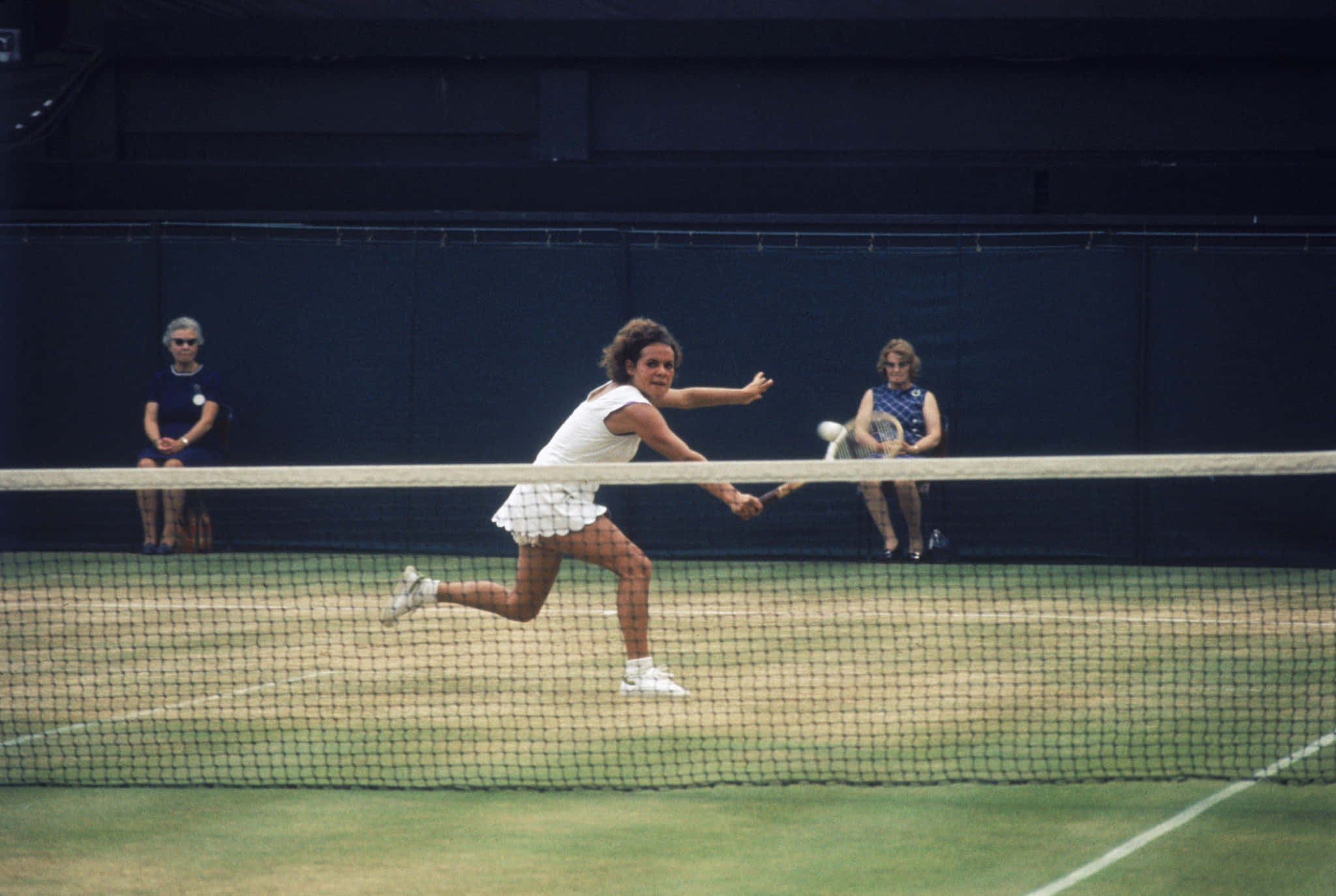 Evonne Goolagong Cawley 後手擊球瞬間 壁紙 — 一名女網球手在草地場上向前衝刺正手擊球，觀眾坐在網後。