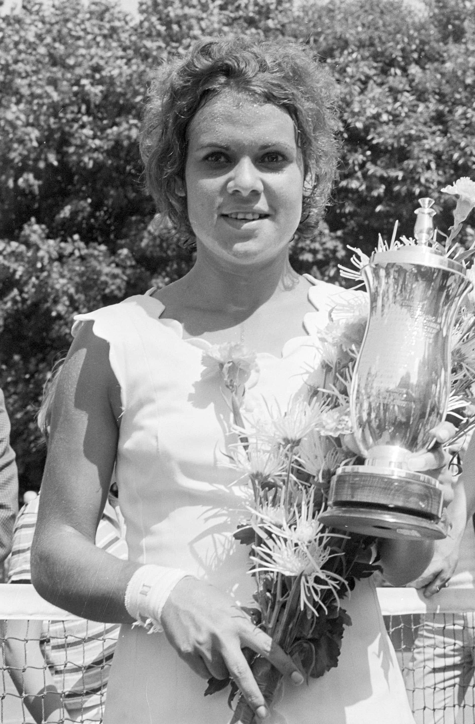 Evonne Goolagong Cawley 與1971年荷蘭公開賽獎盃 壁紙 — 一位微笑的白裙女士在網球場上拿著一個大獎盃和一束鮮花。