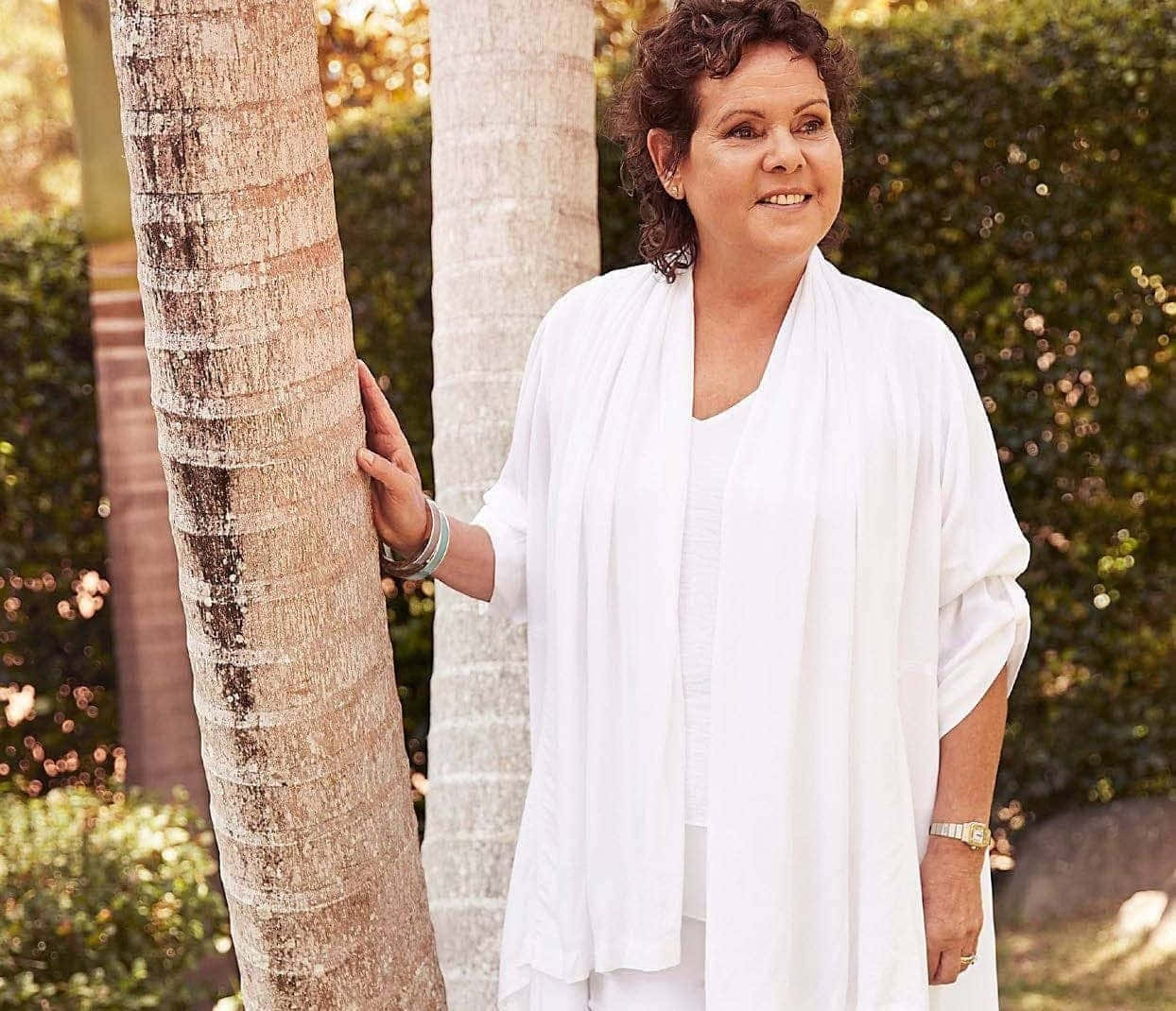 Evonne Goolagong Cawley — 時光永恆的優雅風采 壁紙 — 一位身穿白色服裝的女性在陽光明媚的花園中靠著棕櫚樹，溫柔地微笑。