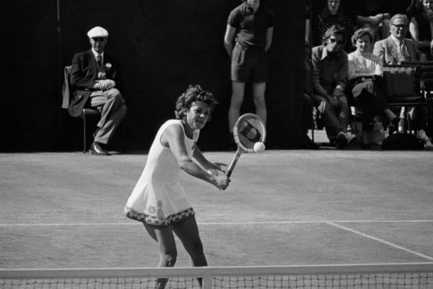 Evonne Goolagong Cawley 澳洲網球選手 壁紙 — 一張復古黑白照片，顯示一位女網球運動員在草地上揮拍，觀眾在旁觀看。