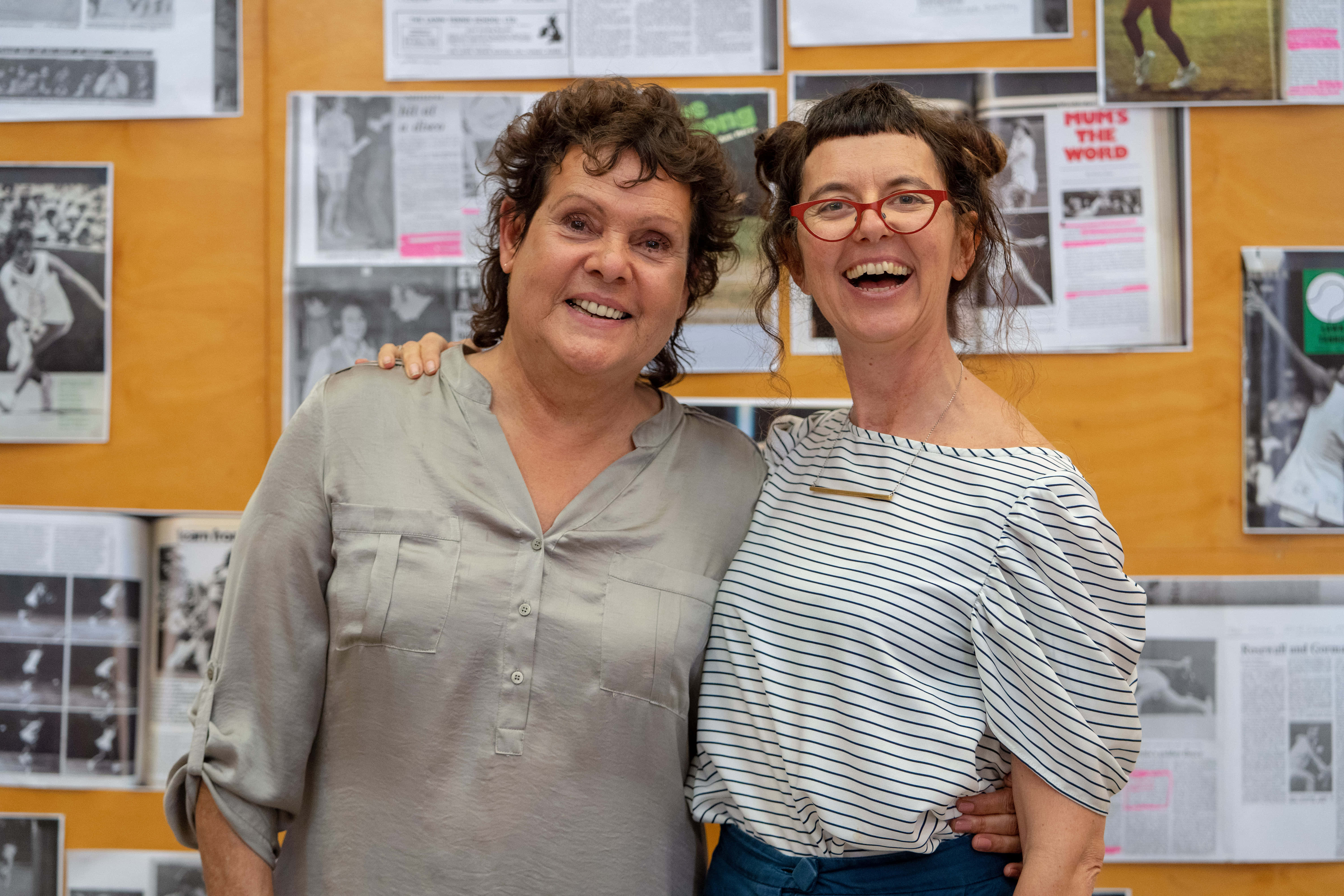Evonne Goolagong Cawley 與 Andrea James 進行坦誠對話 壁紙 — 兩位女士喺鋪滿報紙剪報嘅牆前面企得好近，開心咁大笑。