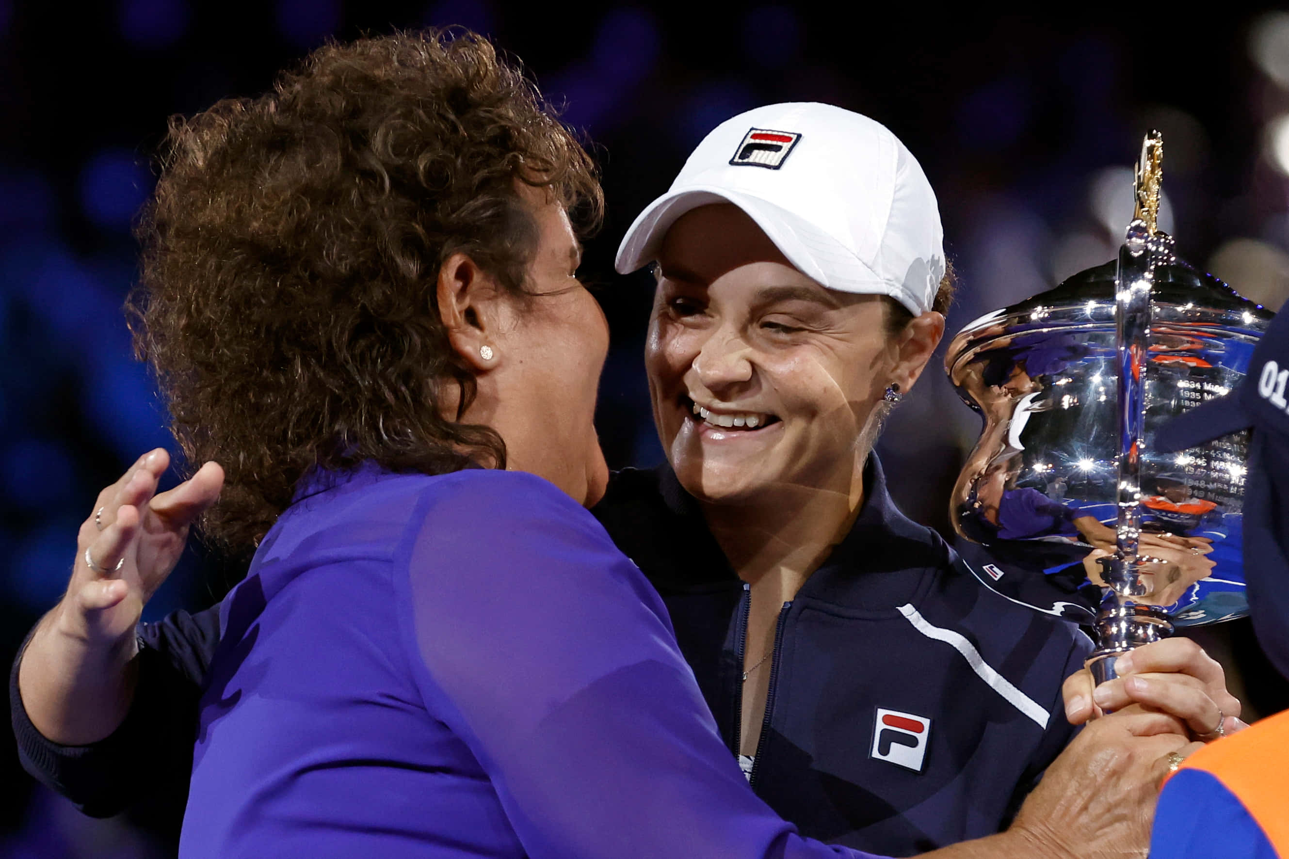 Evonne Goolagong Cawley 為 Ashleigh Barty 道賀 壁紙 — 一位戴白色帽子的微笑網球冠軍，擁抱一位穿紫色外套的女性，同時手持閃亮的銀色獎盃。