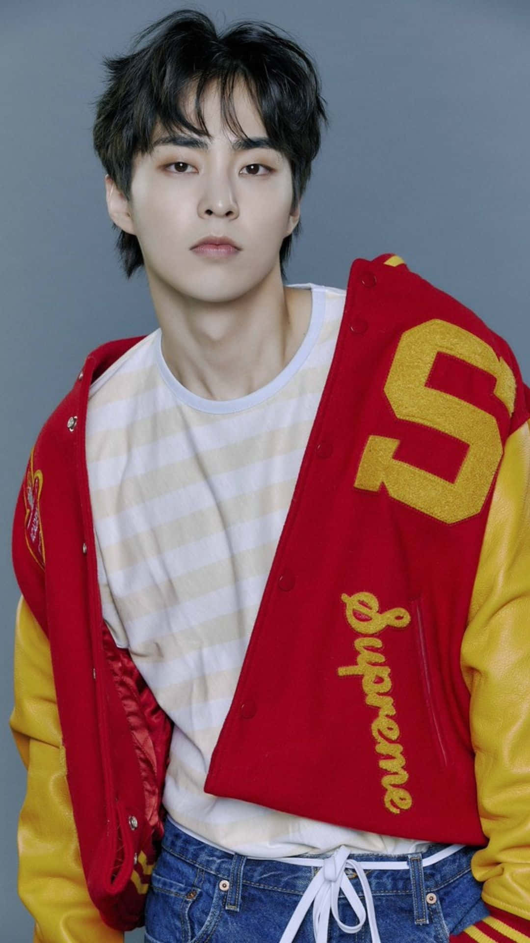 EXO 成員群像合影 壁紙 — 一位黑髮年輕男子身穿紅黃色印有 'Supreme' 品牌的棒球外套，內搭條紋襯衫和牛仔褲，背景為灰色。