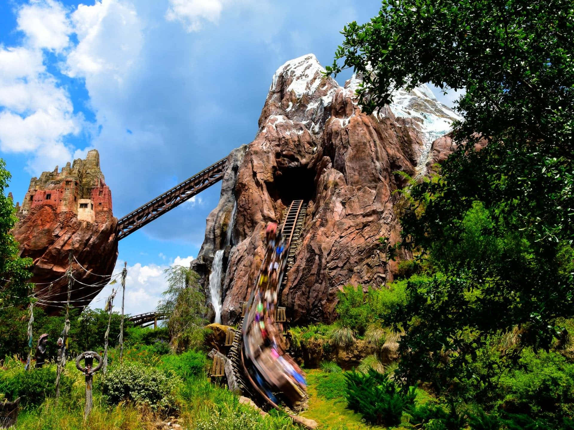 迪士尼世界 Expedition Everest 過山車縮放背景 壁紙 — 一架過山車喺晴朗藍天下，沿住岩石懸崖飛馳，穿過茂密叢林，形成模糊嘅影像。