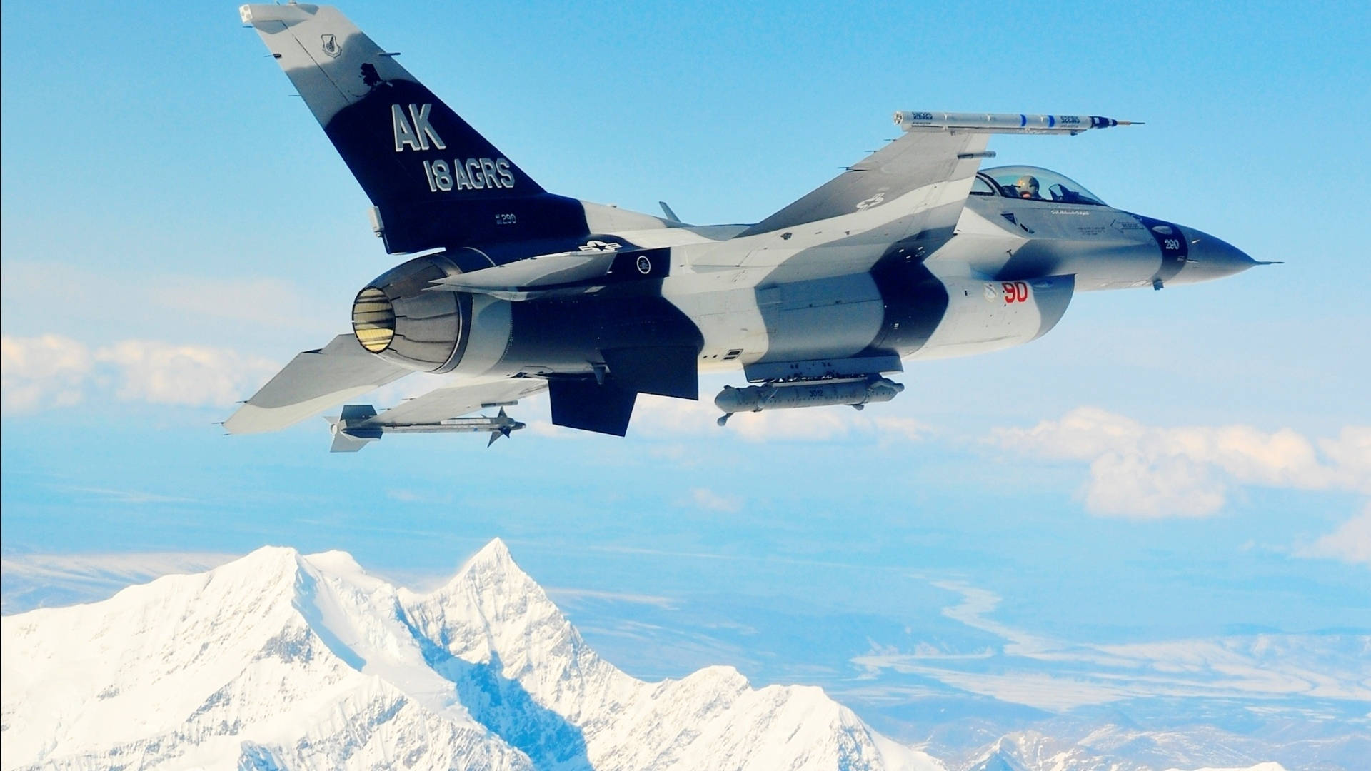 F-16戰隼戰鬥機 壁紙 — 一架灰色戰鬥機在晴朗藍天和零星雲朵下翱翔於積雪山脈之上。