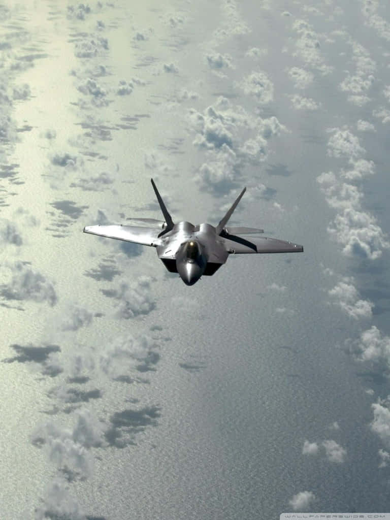 F-22猛禽俯拍Android巨型飛機背景 壁紙 — 一架流線型銀色戰鬥機在明亮的日光下，翱翔於點綴著散落白雲的廣闊海洋之上。