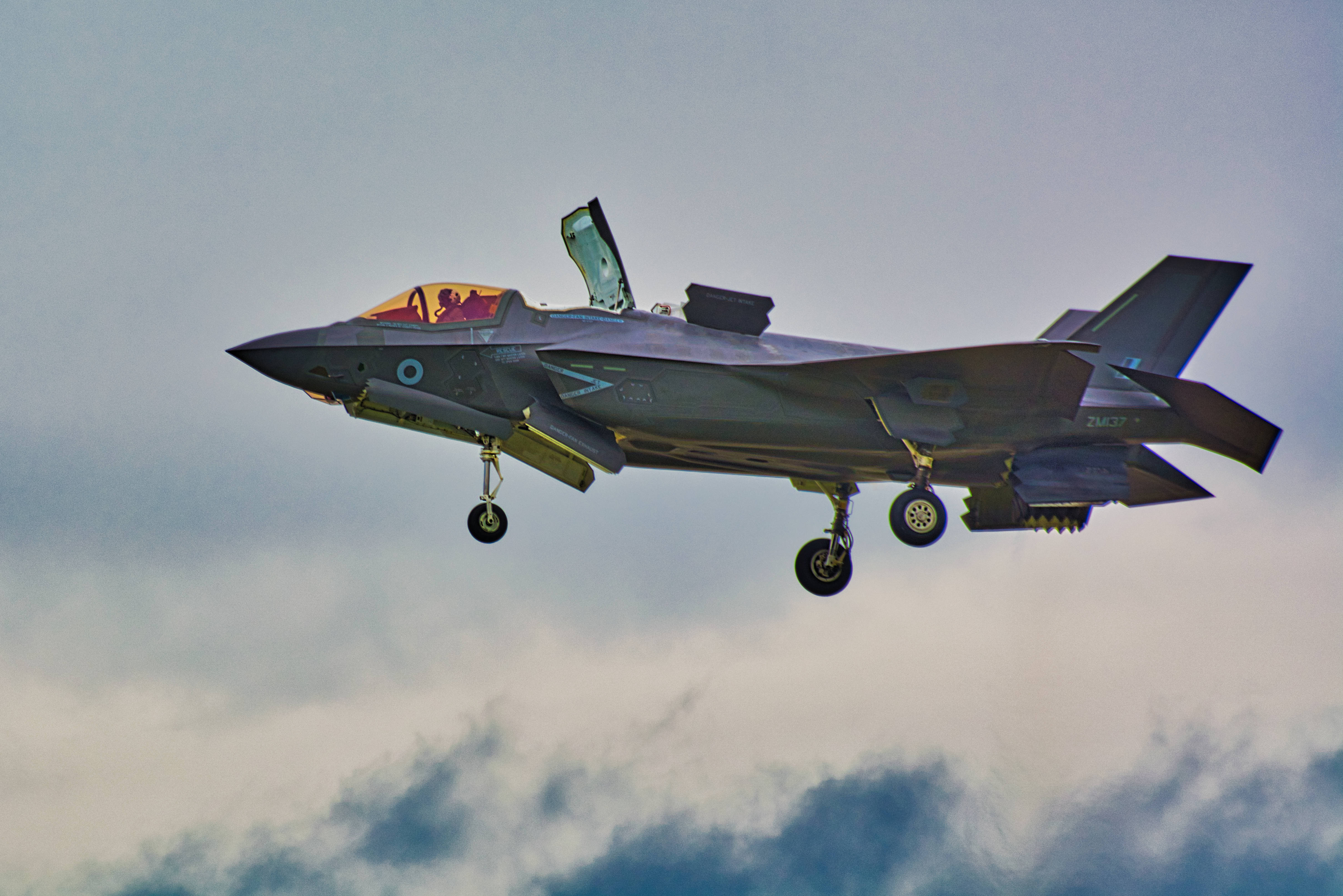 F-35B戰鬥機航空展 壁紙 — 一架灰色戰鬥機放下起落架，打開座艙蓋，在雲層密佈的天空中翱翔，露出裏面的飛行員。