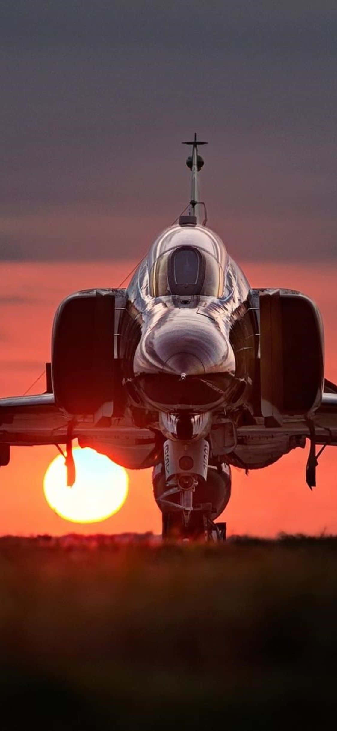 F-4幻影戰機夕陽降落Android巨型飛機背景 壁紙 — 一架流線型戰鬥機在跑道上向前，夕陽在它後面落下，將熾熱的橙色和紅色色調灑滿天空。