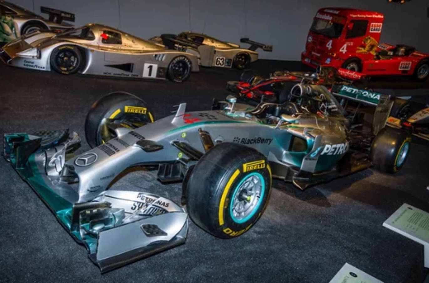 極速之王 - 梅賽德斯F1 4K 壁紙 — 一架流線型銀色和青綠色的 Formula 1 賽車配備 Pirelli 輪胎，在博物館內與其他賽車一同於人造燈光下展出。