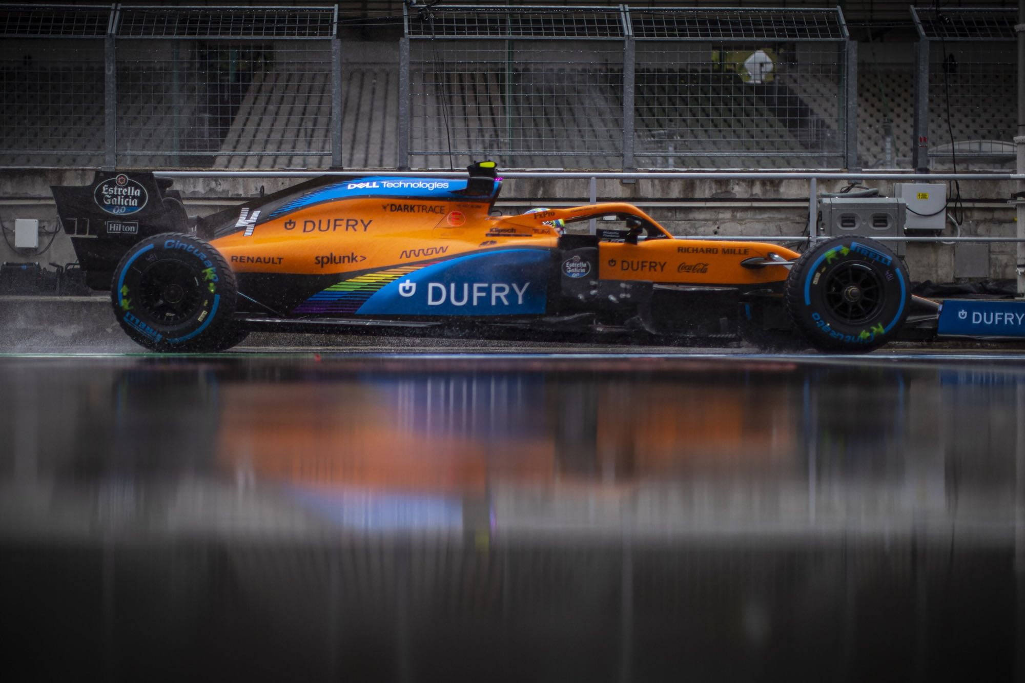 F1 Dufry 麥拉倫 2019 壁紙 — 一輛充滿活力的橙藍色一級方程式賽車在潮濕賽道的水坑中飛馳，倒映其大膽塗裝於陰天之下。