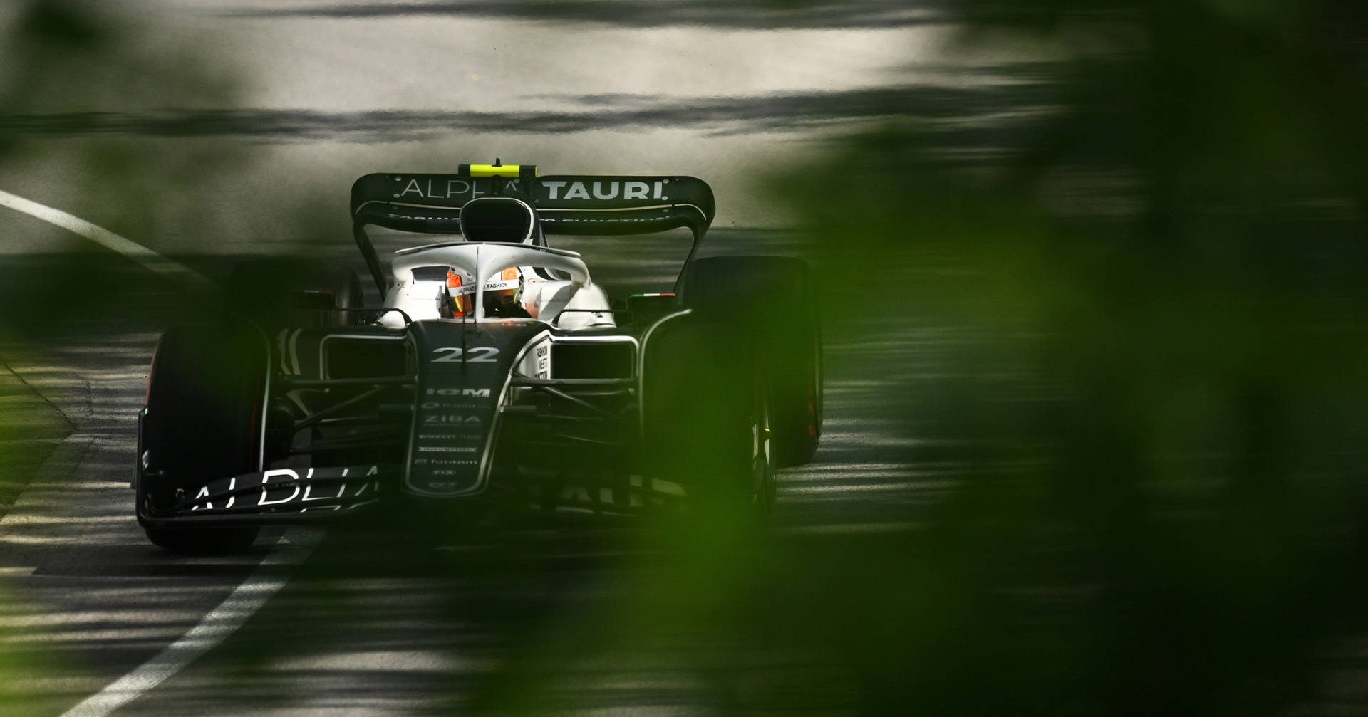 葉子後的F1賽車桌面壁紙 壁紙 — 一架時尚的Alpha Tauri一級方程式賽車在模糊的森林隧道中奔馳，綠色樹葉框住畫面。