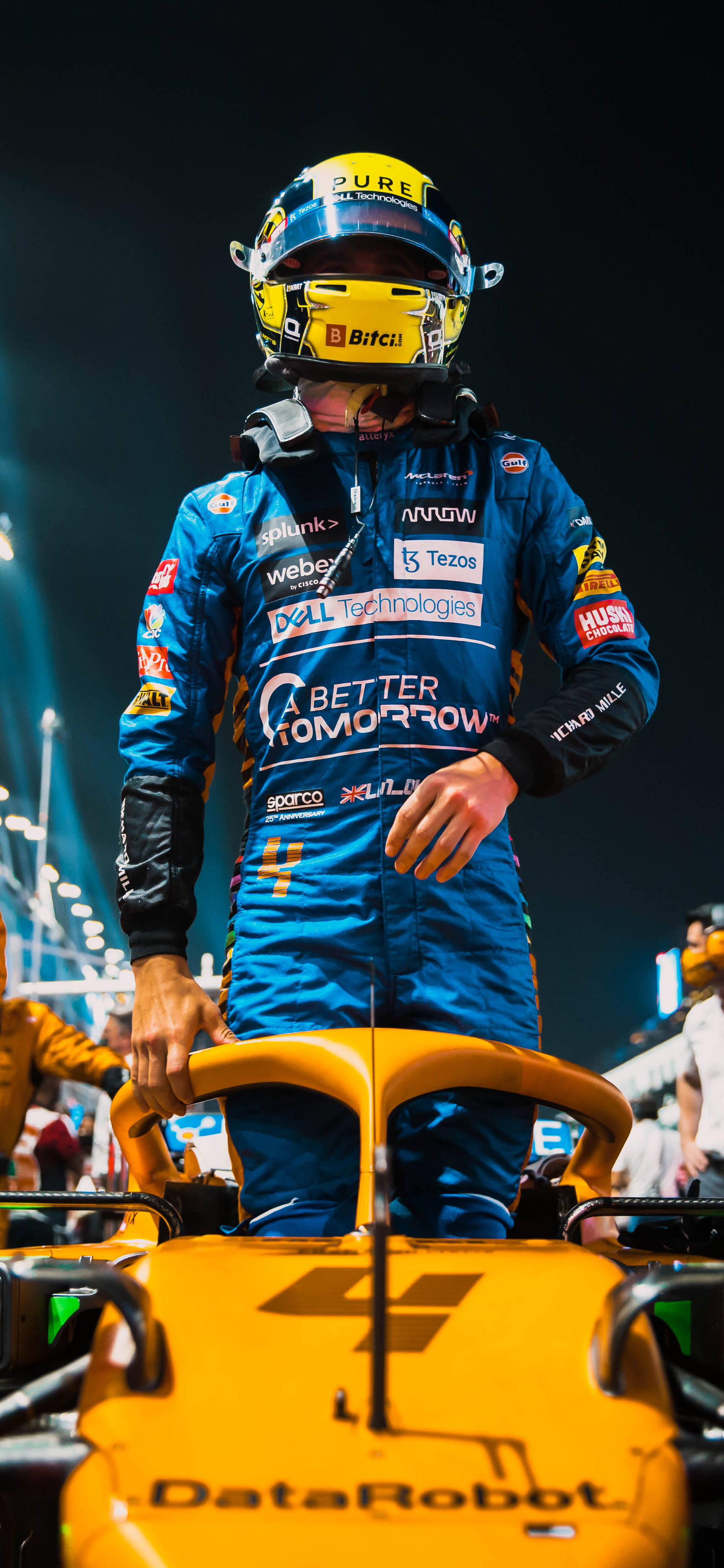 F1 Norris 2021 沙特阿拉伯大獎賽 iPhone 壁紙 — 一位身穿藍色賽車服的F1車手在夜間體育場燈光下站在他亮黃色的賽車上。