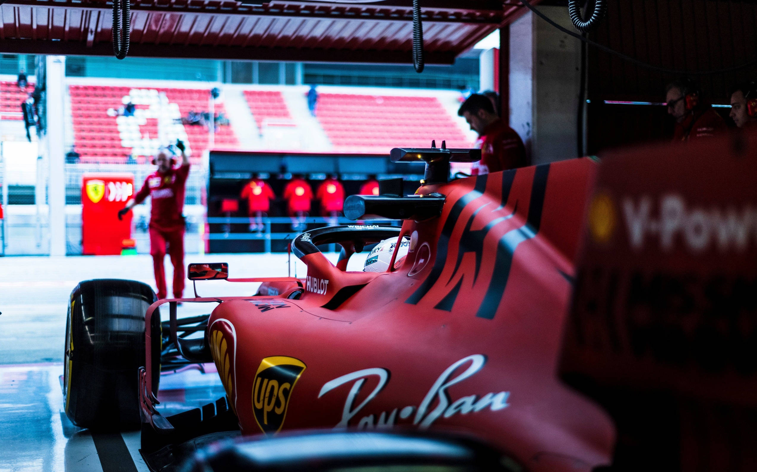 衝向終點線 - F1賽車世界 壁紙 — 一輛紅色法拉利一級方程式賽車在維修站車庫內，背景可見車隊成員和鮮紅色的體育場座位。