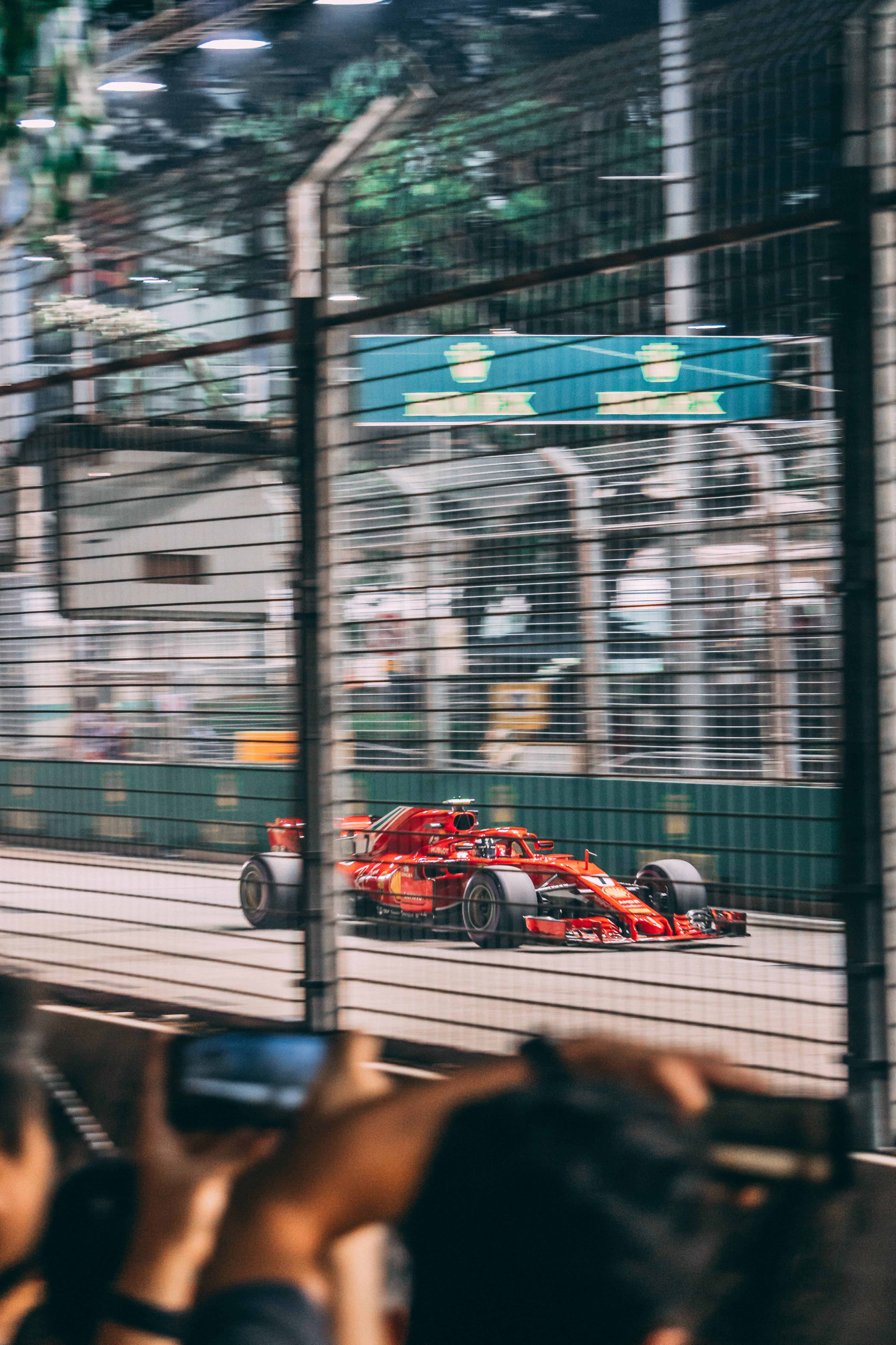 F1 賽車運動聯賽 壁紙 — 一輛紅色一級方程式賽車飛馳經過圍欄，觀眾在明亮的賽道燈光下拍攝比賽。