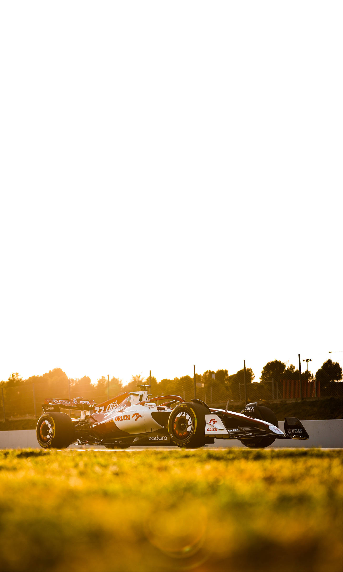 F1 阿爾法·羅密歐 C42 黃昏 iPhone 壁紙 — 一架時尚的白色和橙色賽車在夕陽下沿著賽道滑行，前景是金色的草地。