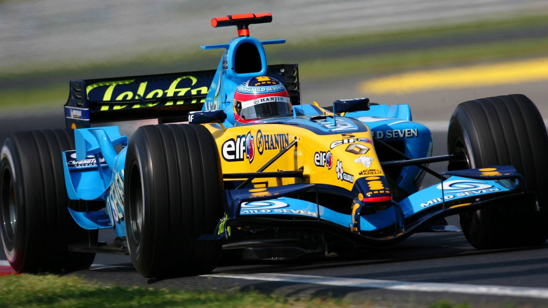 F1 雷諾R25 2005 壁紙 — 一輛藍黃色的一級方程式賽車在賽道上疾馳，背景是模糊的草地和瀝青路面。
