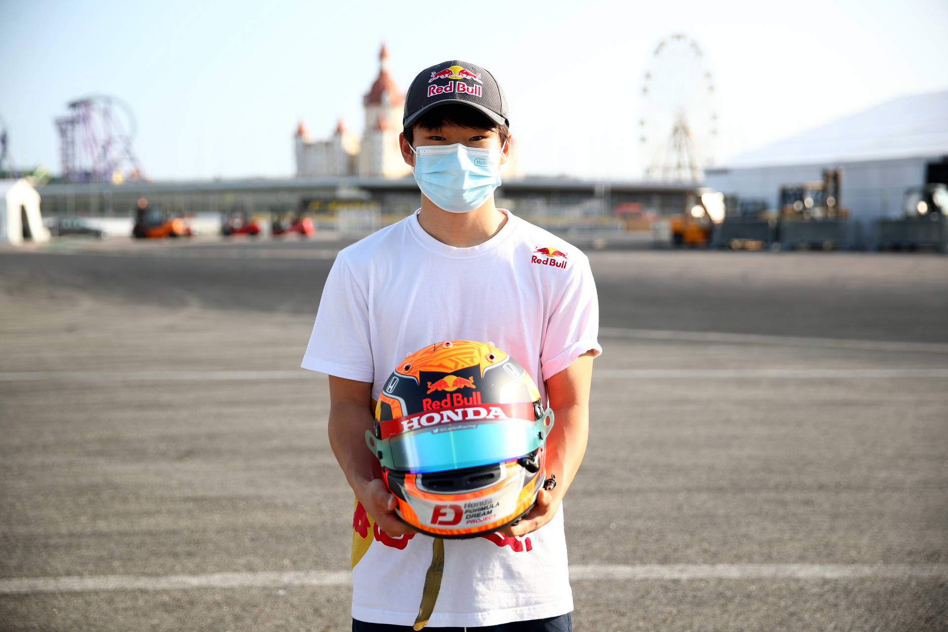 F1 駕駛手 Yuki Tsunoda 戴面罩手持頭盔 壁紙 — 一名戴住Red Bull帽同口罩嘅年輕男子，喺有摩天輪背景嘅賽車場上揸住一個Honda賽車頭盔。