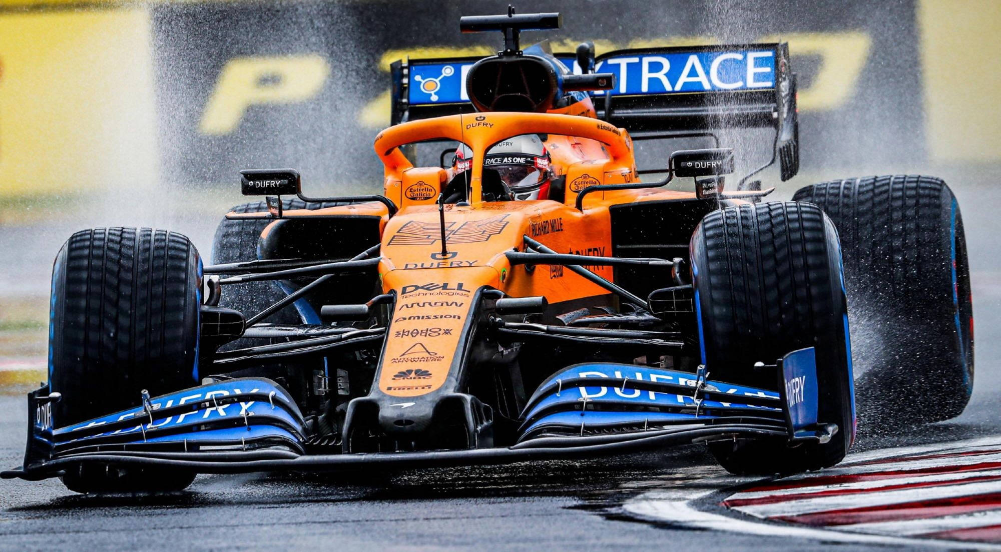 F1 麥拉倫賽車 壁紙 — 一輛橙色和藍色的一級方程式賽車在濕滑的彎道上高速行駛，在潮濕的賽道上濺起水花。