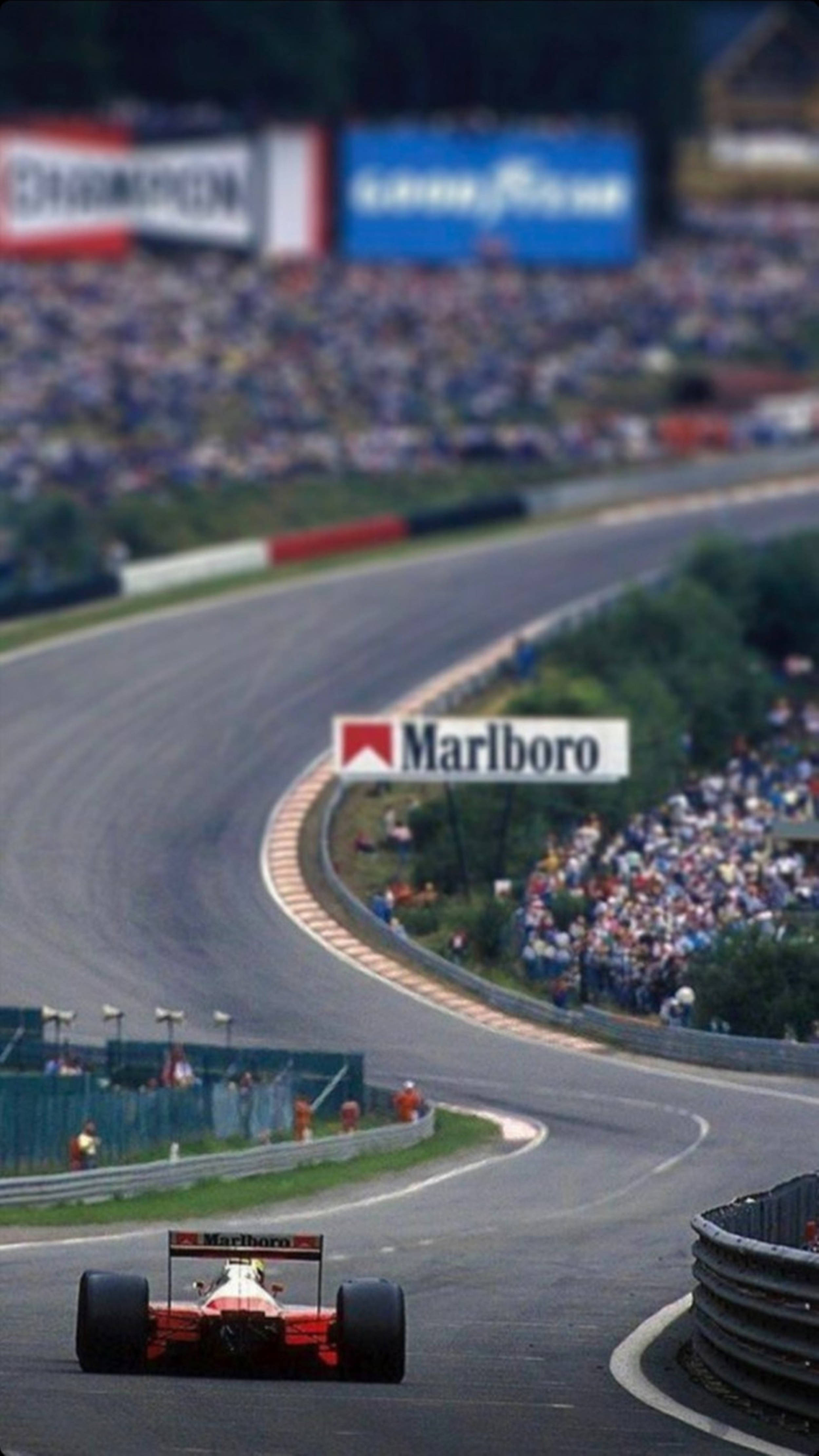 F1手機1988年比利時大獎賽 壁紙 — 一輛紅色一級方程式賽車在陽光明媚的賽道上高速過急彎，背景是歡呼的人群和萬寶路橫幅。