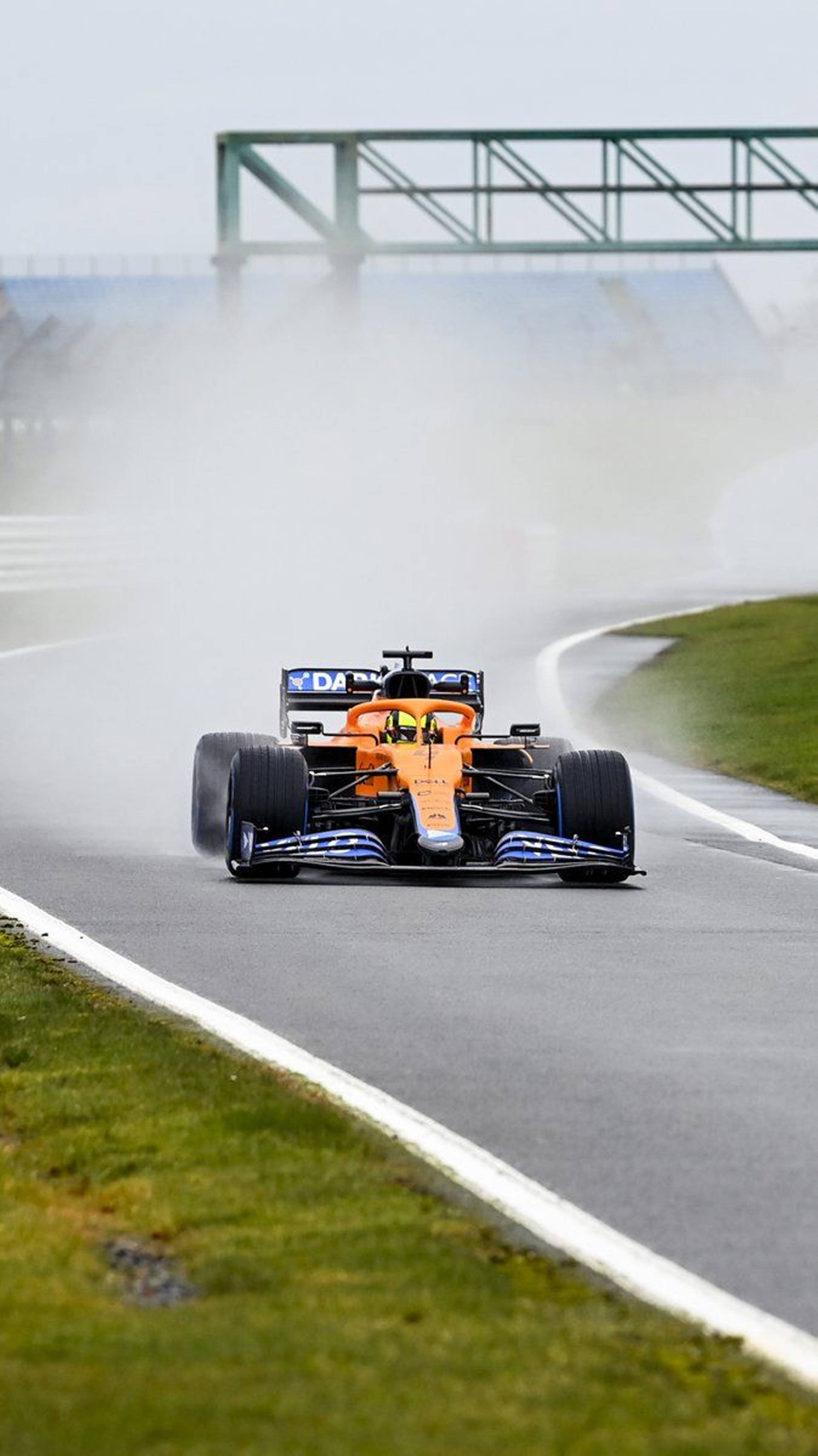 F1手機麥拉倫MCL35煙霧效果 壁紙 — 一輛橙色和藍色的F1賽車在陰天之下，於潮濕賽道的彎角高速飛馳，激起水霧。