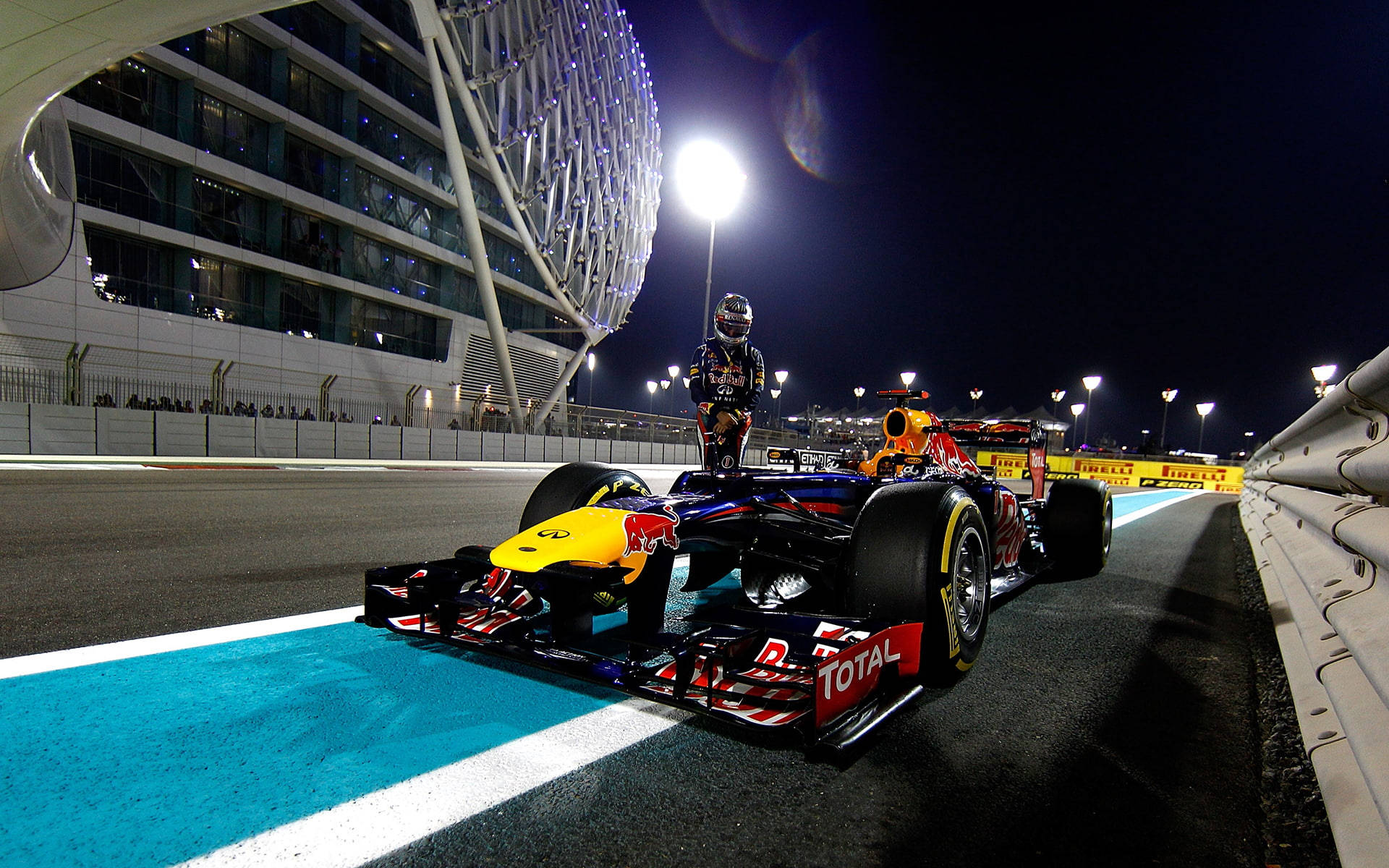 F1大獎賽2018精彩瞬間 壁紙 — 一輛紅牛一級方程式賽車與車手在夜間準備就緒，停在明亮照明的賽道上，旁邊是一座未來主義的體育場。