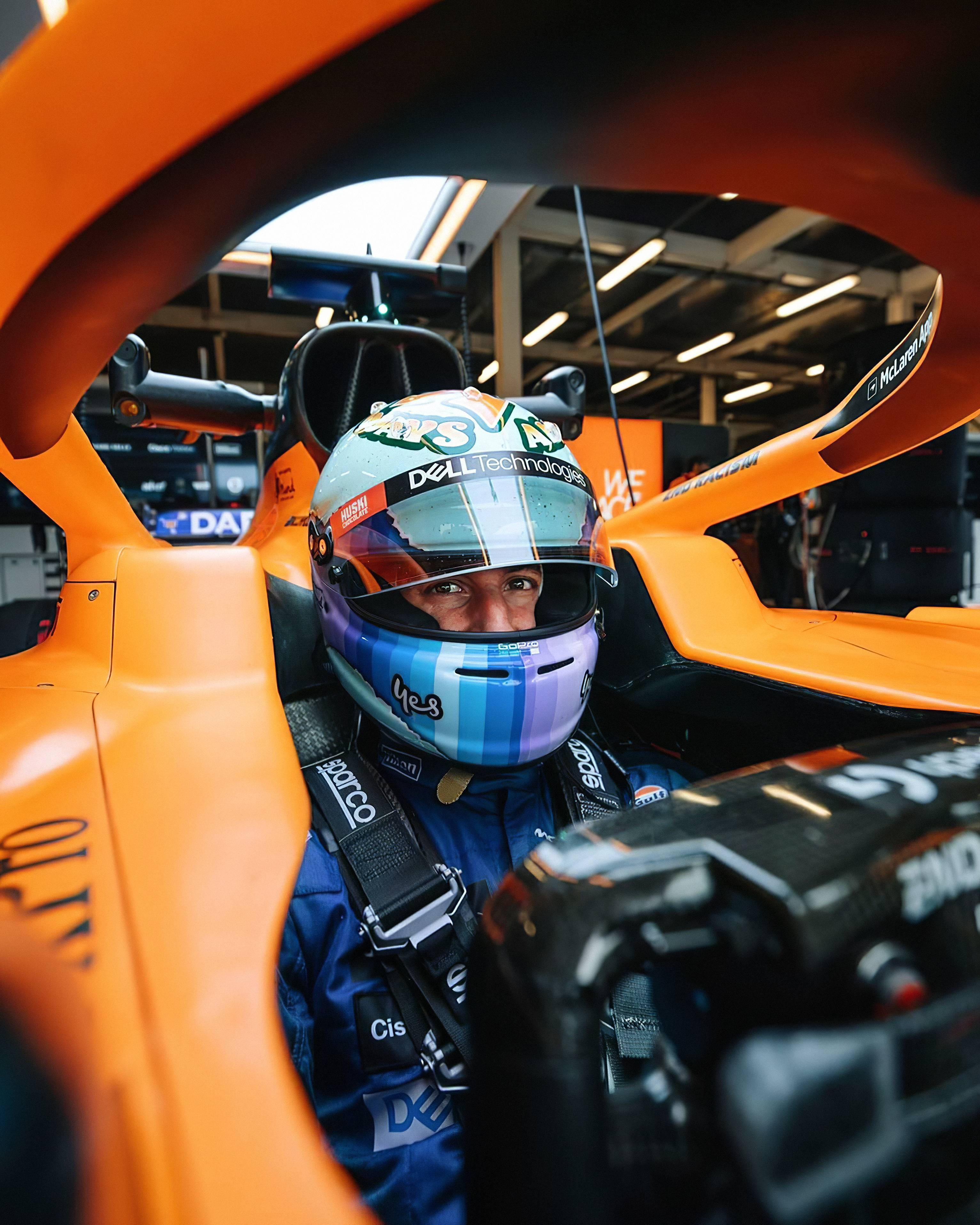 F1明星丹尼爾·里卡多戴頭盔微笑 壁紙 — 一級方程式車手坐在充滿活力的橙色麥拿倫賽車駕駛艙內，戴著帶有贊助商標誌的彩色頭盔。