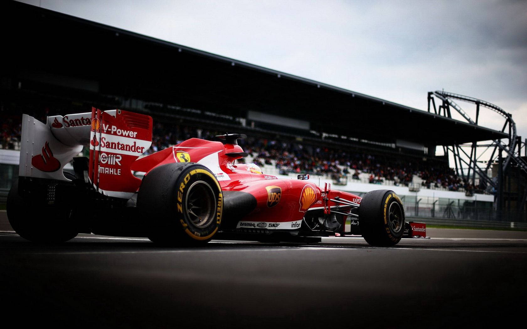 F1賽車桌面紅色美學 壁紙 — 一輛紅色法拉利一級方程式賽車在陰天之下沿著賽道飛馳，背景是坐滿觀眾的看台。