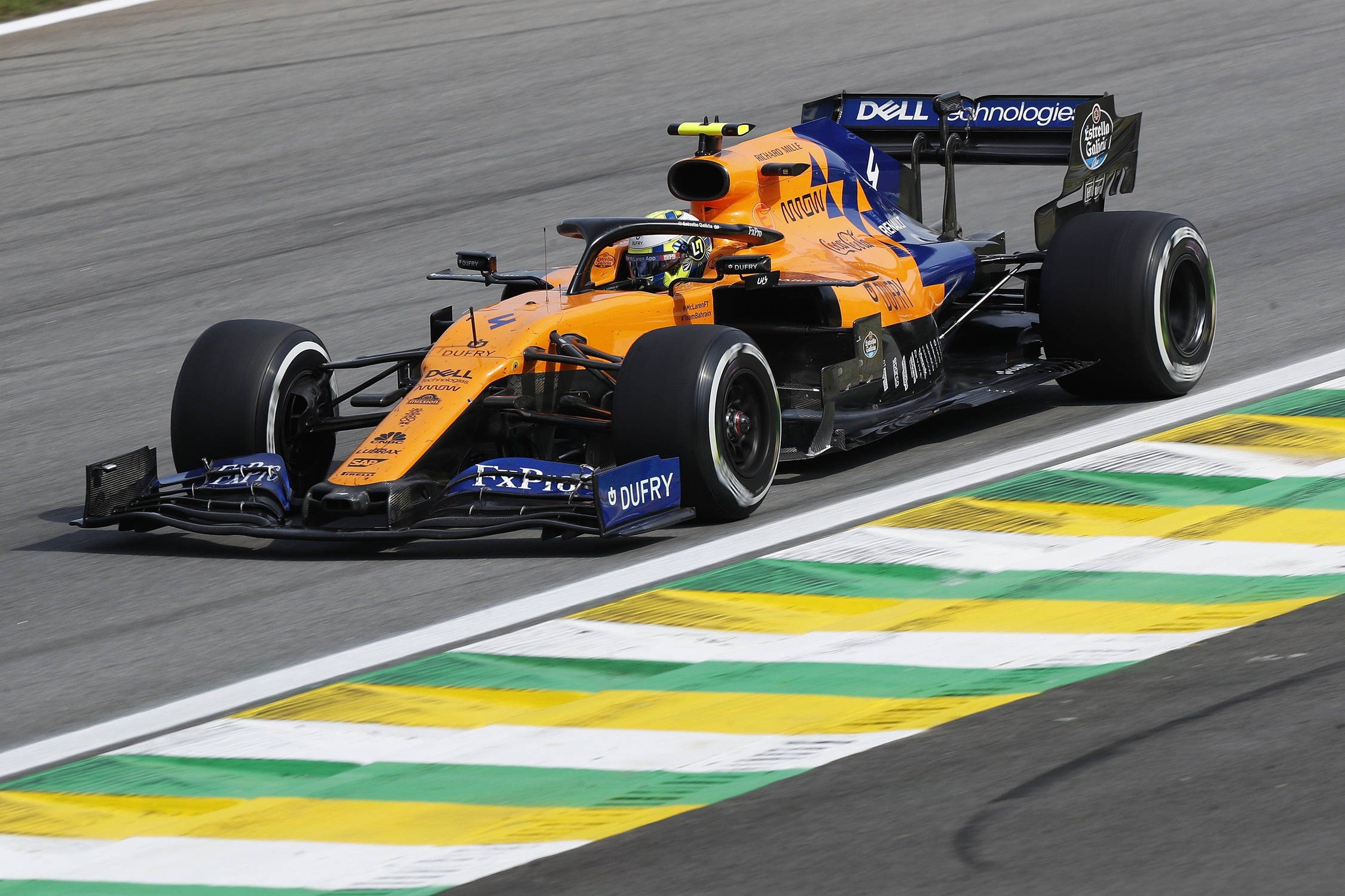 F1賽車極速競技桌面場景 壁紙 — 一輛充滿活力的橙藍色一級方程式賽車在明亮的日光下，沿著設有綠、黃、白色路緣的賽道疾馳。