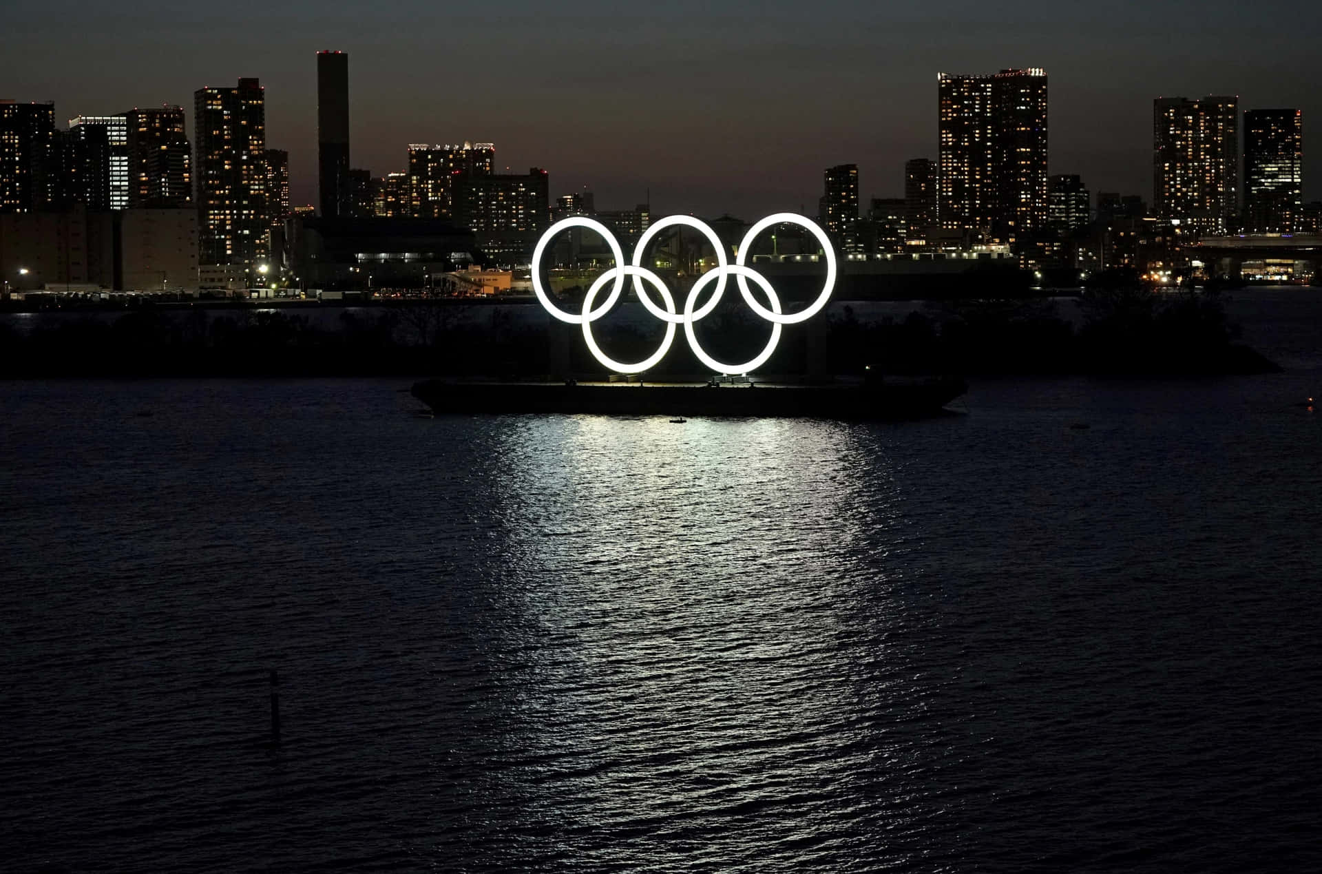 奧運五環在夜間的水面上發光，背景是城市燈光。