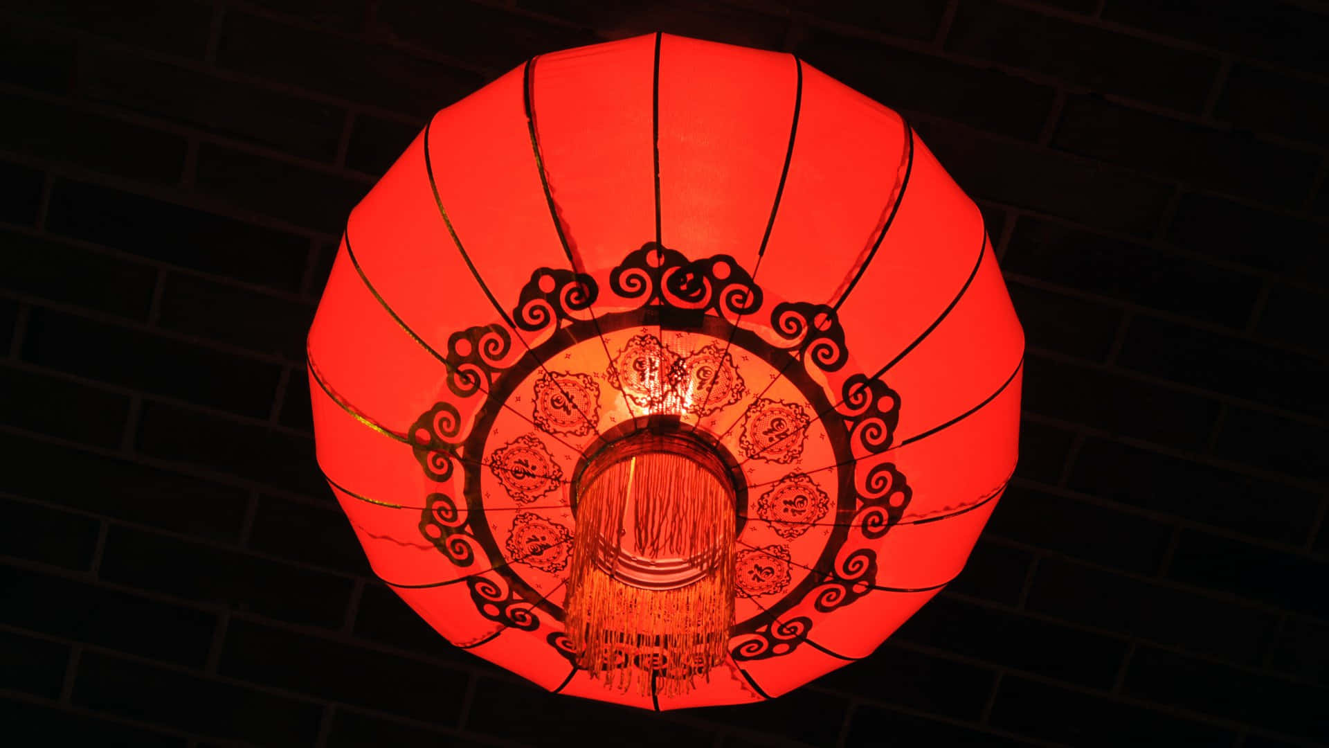  壁紙 — 一個發光的紅色紙燈籠，帶有複雜的黑色捲曲花紋，夜晚掛在深色磚牆上。