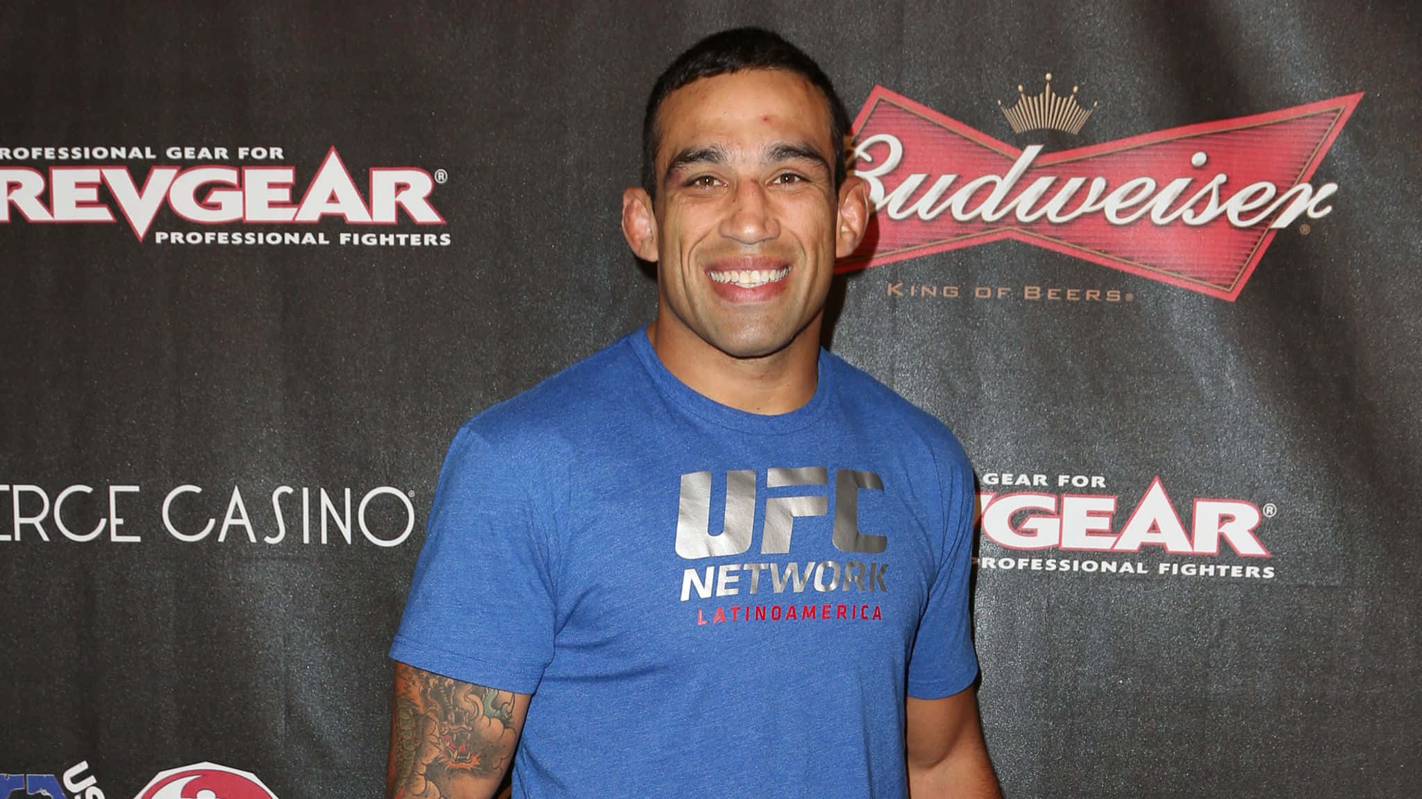 Fabricio Werdum 出席 BAMMA USA 活動 壁紙 — 一名微笑的男子身穿藍色UFC Network T恤，站在印有Budweiser和RevGear標誌的品牌背景前。