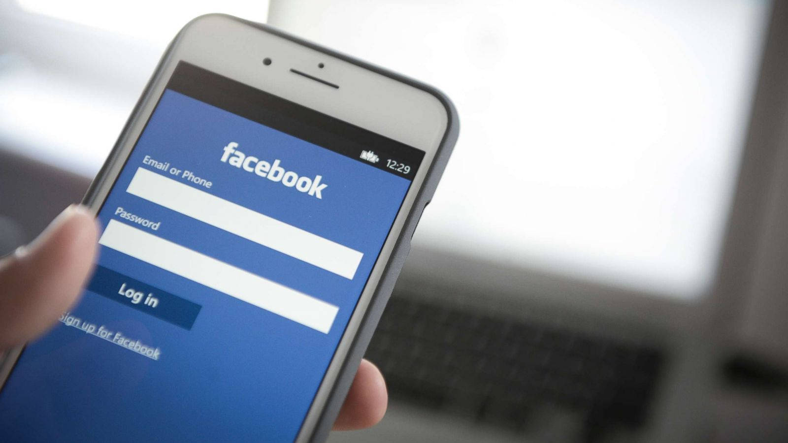 Facebook 移動登入介面 壁紙 — 一隻手拿著智能手機，顯示Facebook登入畫面，背景有模糊的筆記型電腦。